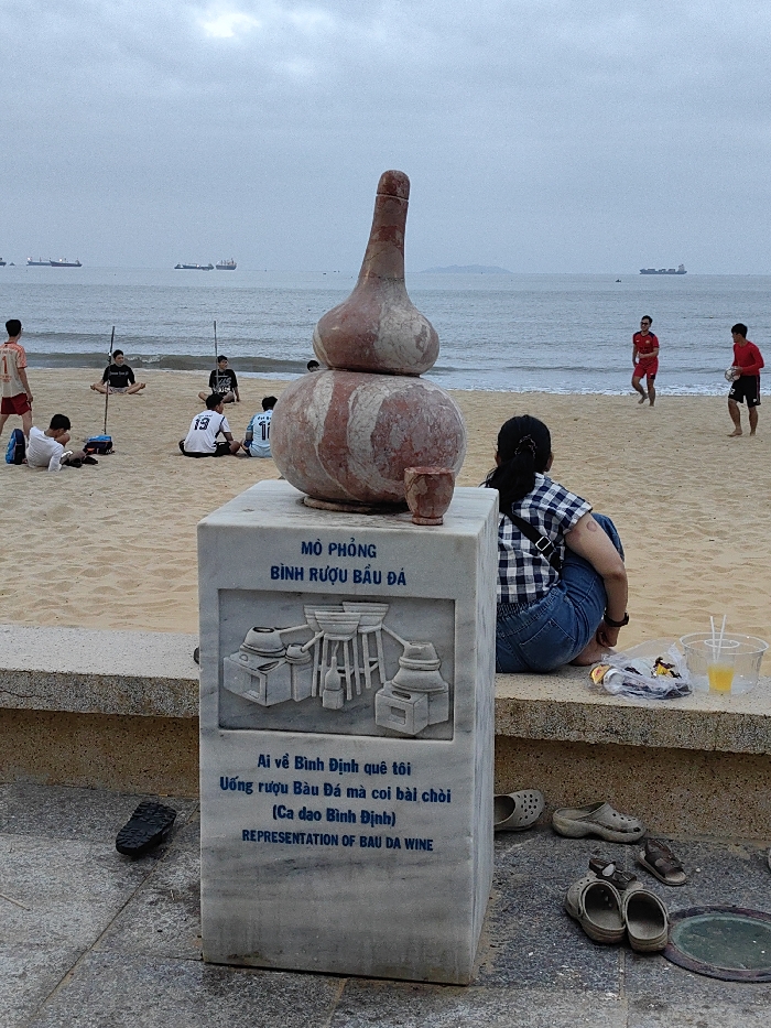 The scene depicts a beachfront area in Quy Nhon, Vietnam, under an overcast sky. In the foreground, a gray marble monument stands on a tiled surface, topped by a reddish-brown, two-tiered gourd-shaped sculpture with a smaller, matching cup beside it. The monument's front panel is engraved with Vietnamese text, including "MÔ PHỎNG BÌNH RƯỢU BẦU ĐÁ" (Representation of Bau Da Wine), an illustration of traditional brewing apparatus, and a Vietnamese folk poem, followed by "REPRESENTATION OF BAU DA WINE" in English. To the left of the monument, a single black flip-flop rests on the tile. To the right, a person with dark hair, wearing a blue and white checkered shirt and blue jeans, is seated on a concrete ledge, facing the beach with their back to the camera. Adjacent to them on the ledge are a clear plastic cup with a yellow beverage and two straws, and a clear plastic bag containing miscellaneous items. On the ground by the ledge, multiple pairs of footwear, including two pairs of Crocs and brown sandals, are scattered. The midground features a sandy beach where approximately eight individuals are visible. On the left side of the beach, three young men are seated on the sand; one wears a white jersey with the number "13," another a dark shirt, and a third a light jersey with "11." Two slender poles are upright in the sand nearby. Towards the right, two men in red shirts and black shorts are at the water's edge; one walks towards the ocean, while the other holds a light-colored object. The background consists of the ocean under a uniformly gray sky, with several large cargo ships visible on the distant horizon and a faint landmass to the far right.