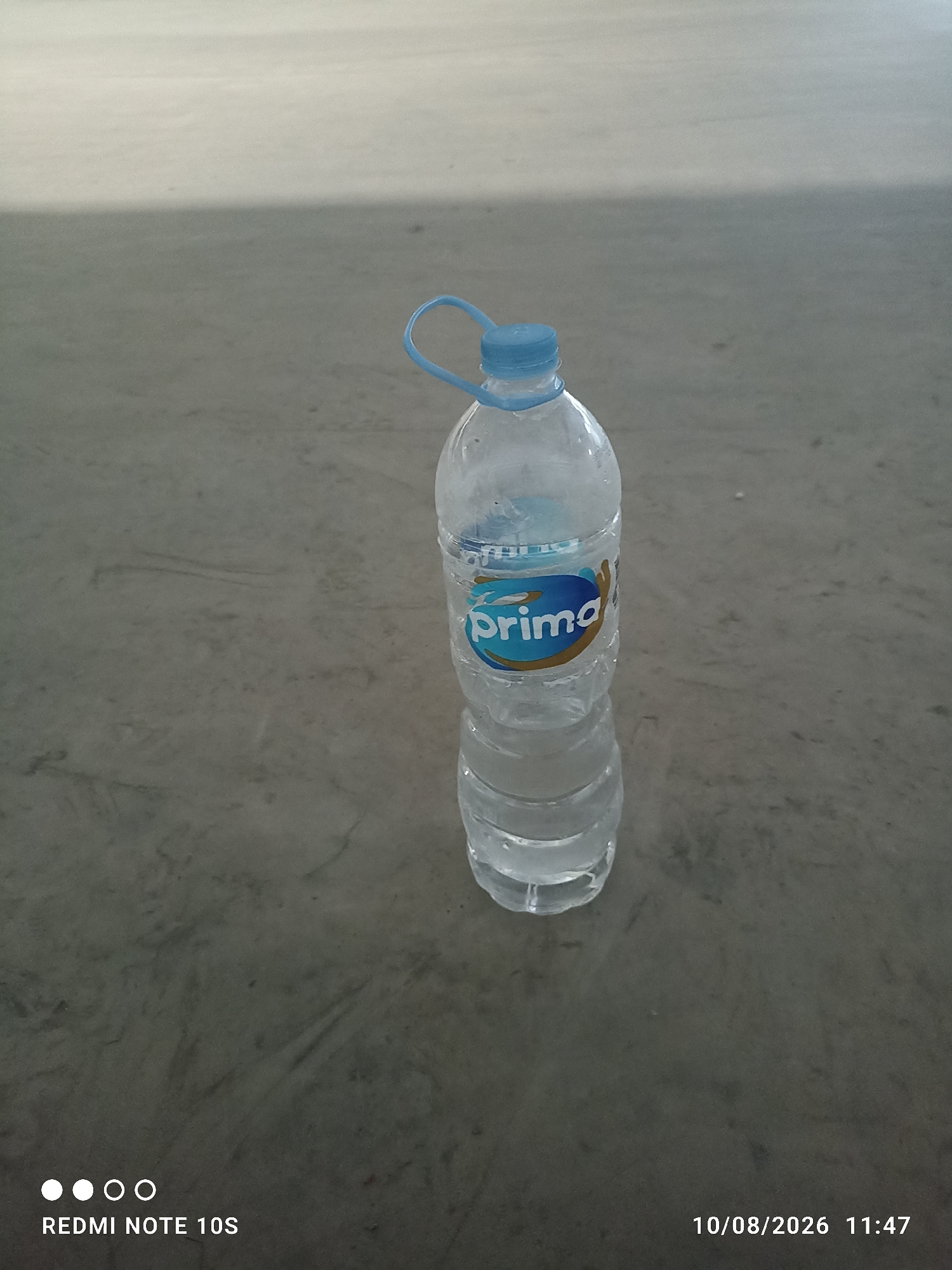 A plastic bottle of Prima brand water sits on a concrete surface in Kosambi Barat, Indonesia. The bottle is partially filled with water and has a blue cap and a blue and gold label. The surface appears to be an outdoor area, possibly a sidewalk or a public space, with some scuff marks and a light shadow cast from above. The image also includes a timestamp of "10/08/2026 11:47" and the text "REDMI NOTE 10S" in the lower left corner.