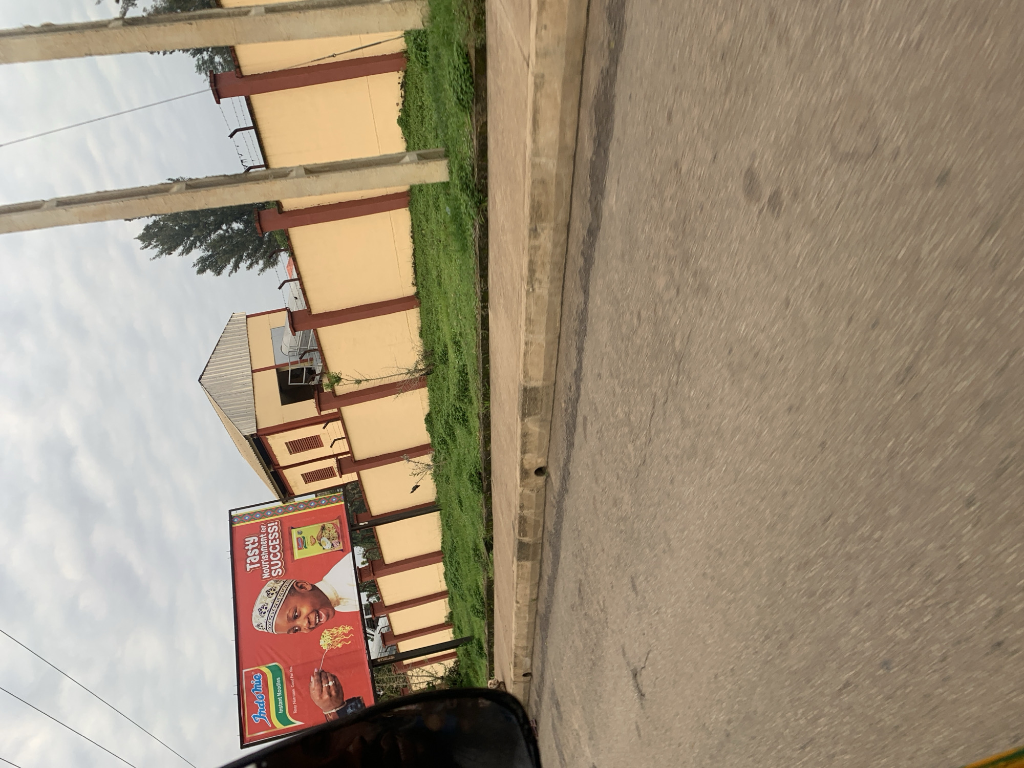 The image captures a street scene in Rafin Par, Nigeria. A large billboard advertising "Indomie Instant Noodles" is prominently displayed. The billboard features a young boy wearing a cap, smiling as he is being fed noodles from a fork. Text on the billboard reads "Tasty Nourishment for SUCCESS!" and "Tasty Nutrition. Good For You." Behind the billboard, a building with a light-colored facade and a corrugated metal roof is visible, bordered by a tan wall with red vertical accents and topped with barbed wire. The wall runs along a grassy verge and a paved sidewalk. The street in the foreground is asphalt. The sky is overcast with visible clouds. The perspective suggests the photo was taken from a moving vehicle.