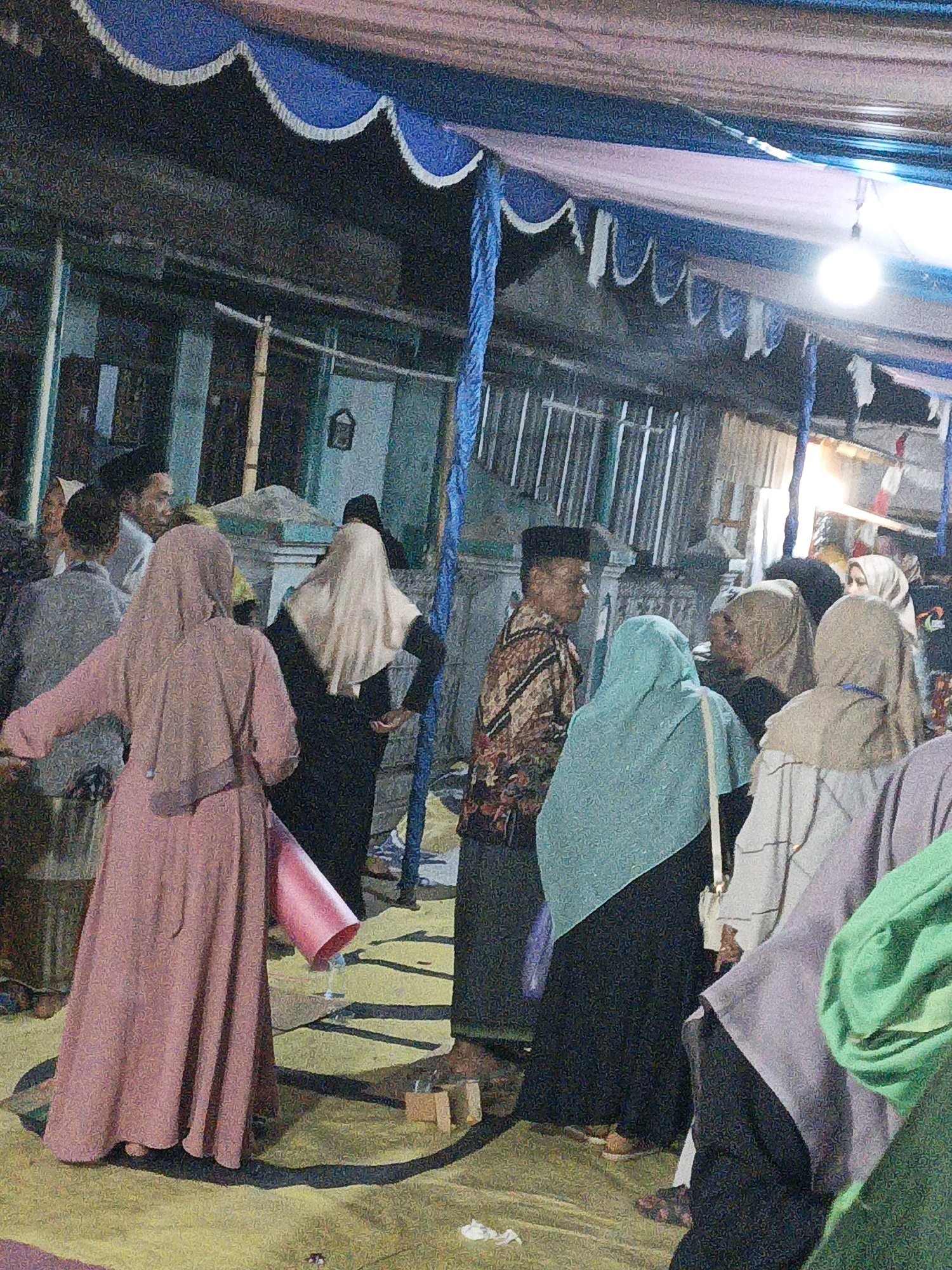 The image depicts an outdoor gathering at night in what appears to be a temporary structure, possibly a tent or canopy, set up in Tanahbaya, Indonesia. The structure is adorned with blue and white fabric draping. Several individuals, mostly women wearing hijabs of various colors, are visible. One woman in the foreground wears a pink hijab and a long pink dress. Another woman next to her wears a black dress and a light-colored hijab. A man in the mid-ground wears a black cap and a patterned shirt. The ground is covered with a yellow mat or carpet, and shadows suggest overhead lighting from a bulb visible in the upper right of the frame. A pink cone-shaped object is held by the woman in the pink dress, and other small objects are scattered on the ground. The background shows a building with corrugated metal fencing. The overall scene suggests a social or religious event taking place after dark.