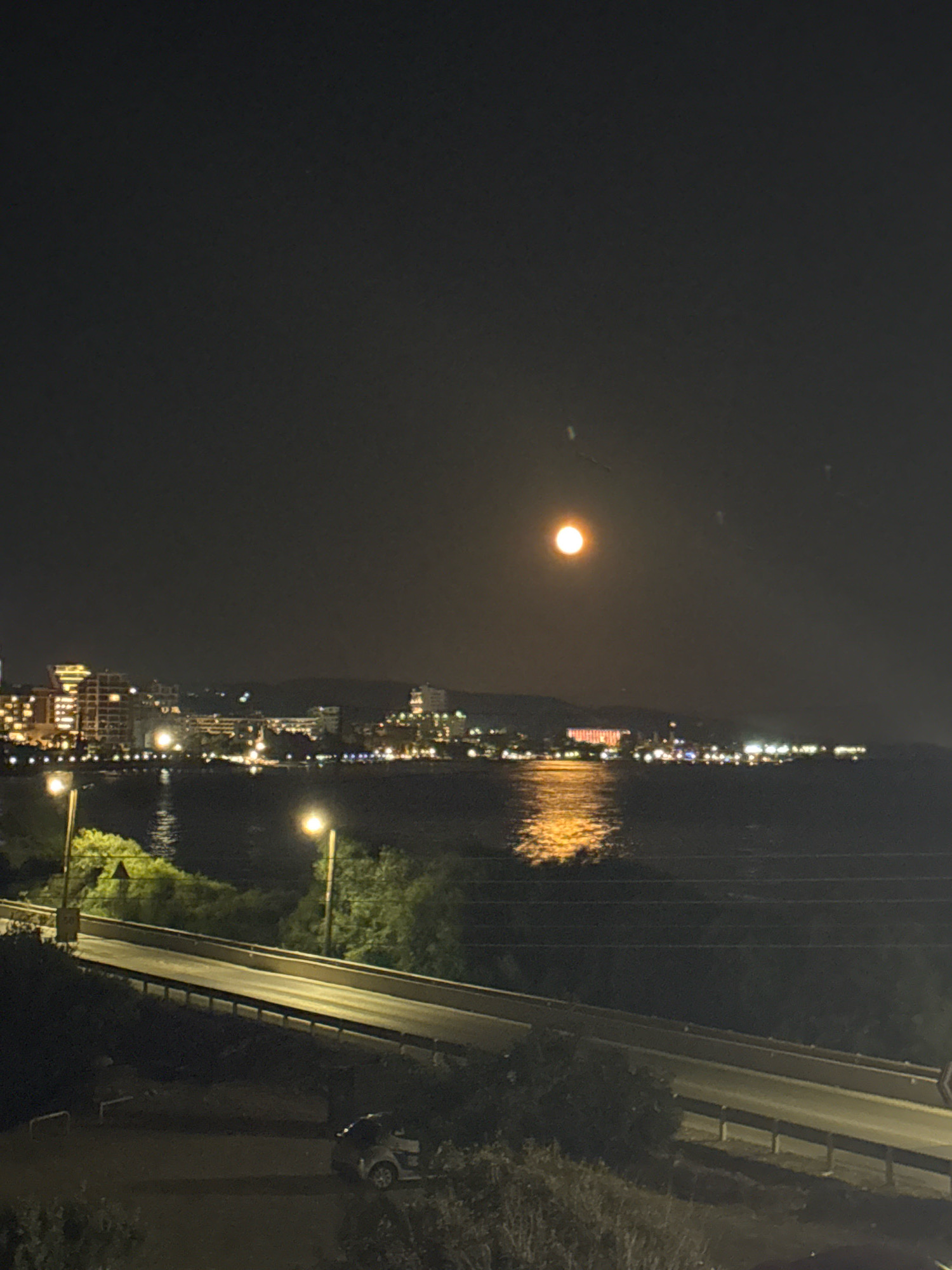 A full moon shines brightly in a dark night sky over the city of Agios Tychona Community, Cyprus. The city lights are visible along the coastline, reflecting on the water. In the foreground, a road winds its way through lush greenery. A small car is parked on the side of the road. The overall scene is serene and peaceful, with the moon and city lights casting a warm glow on the landscape.