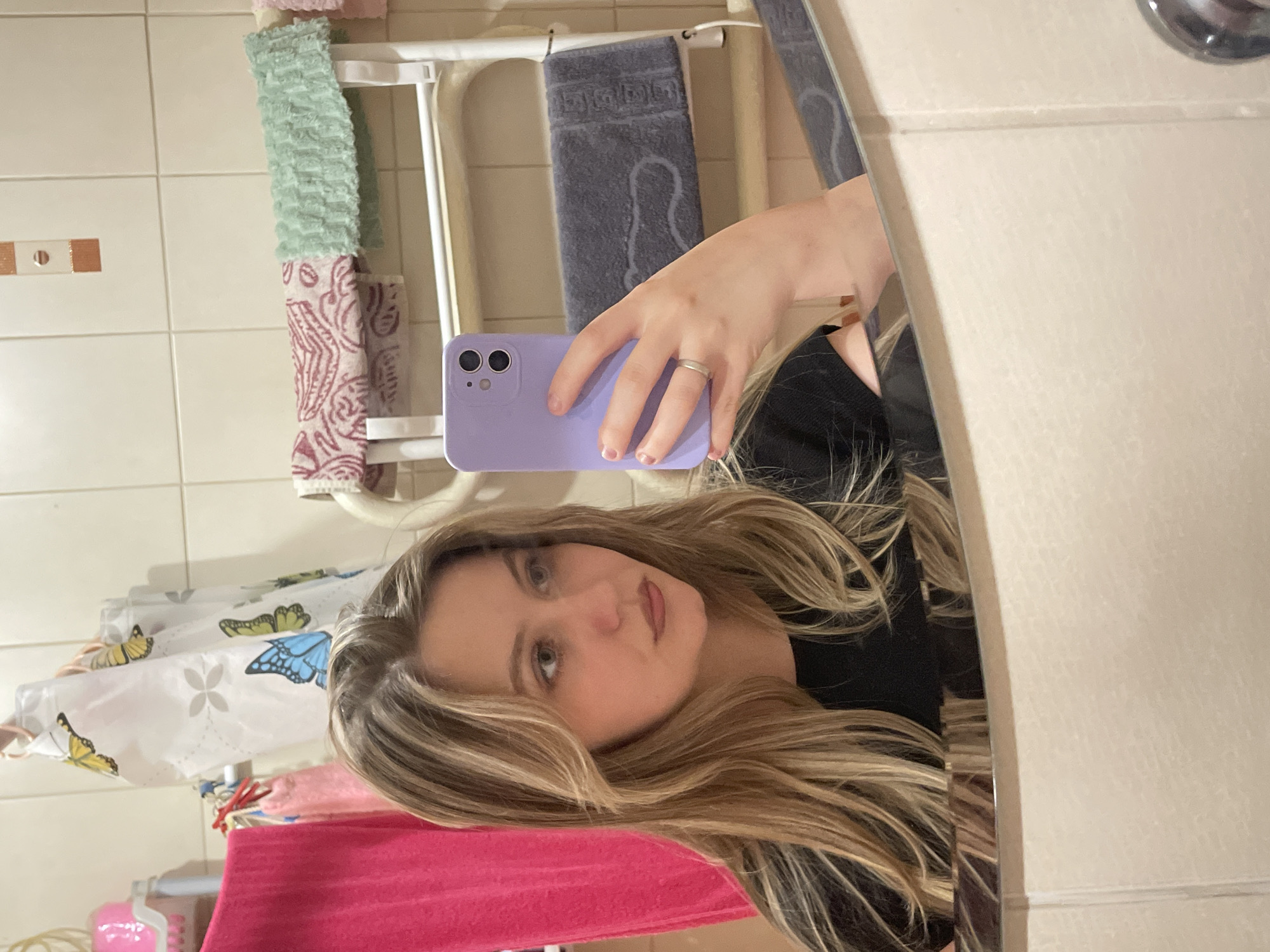 A young woman with blonde hair tied back is taking a selfie in a bathroom mirror. She is wearing a black top and holding a purple phone in her right hand, which is resting on the mirror. The background shows tiled walls, a towel rack with several towels, and a pink basket. The lighting is dim, suggesting it is nighttime or the room is not brightly lit.