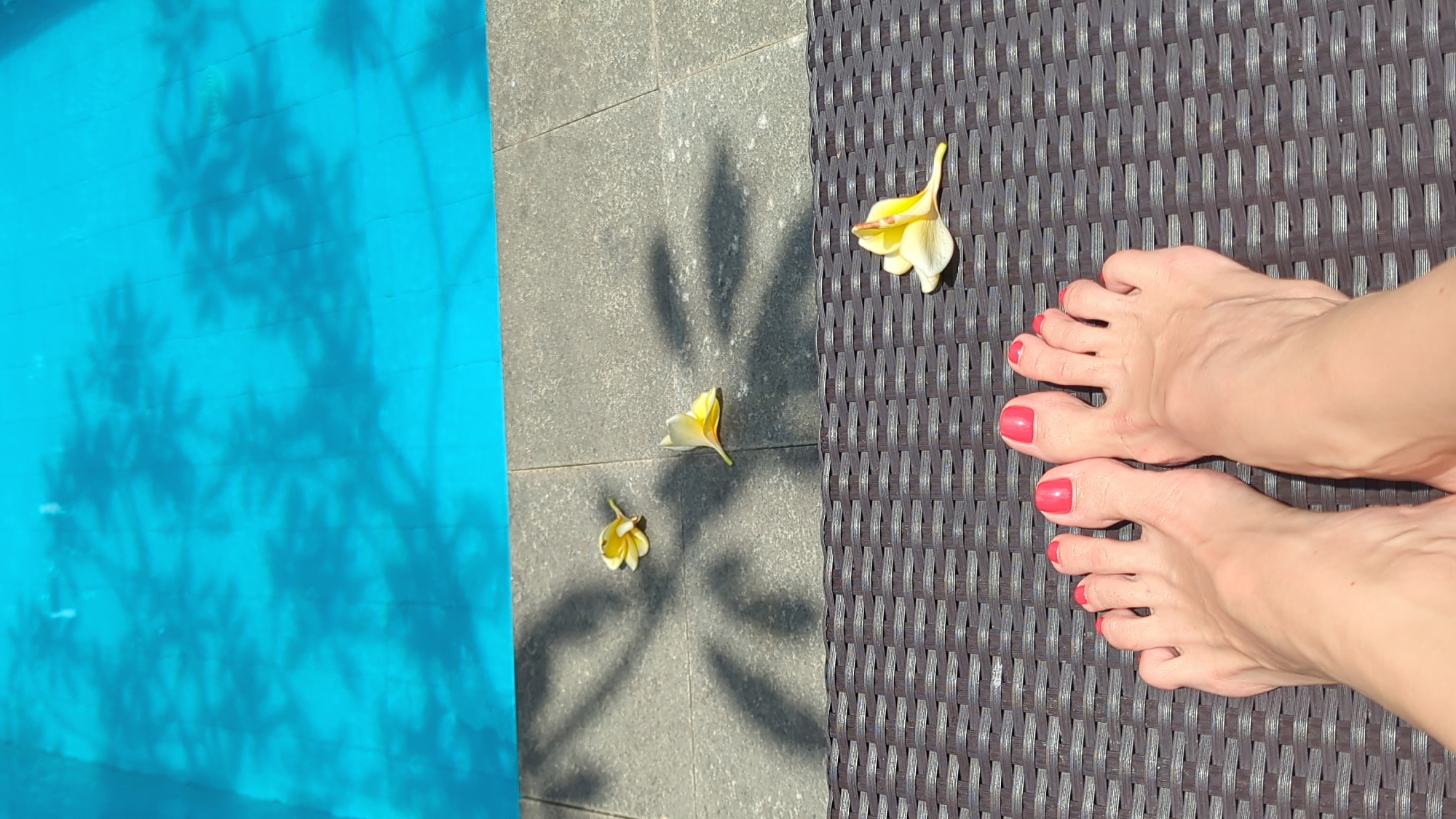 The photograph shows a view of a sunlit poolside area, likely in North Kuta, Indonesia. On the left is a section of a turquoise blue swimming pool, overlaid with shadows suggesting overhanging foliage. The central area consists of grey tiles displaying the shadow of a plant, with three fallen yellow and white frangipani flowers scattered nearby. The right side of the image features a dark brown woven sun lounger on which a pair of feet rest, the toes painted with bright pink nail polish.