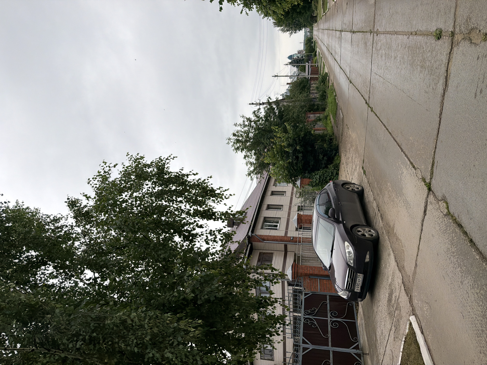 The image depicts a residential street scene in Chaikovsky, Russia, on an overcast day. A dark-colored sedan is parked on the side of a paved road that stretches into the distance, lined by trees and houses. The house in the foreground is a multi-story building with light-colored siding and a brown roof. A decorative metal gate is visible in front of the property. The road itself is made of concrete slabs with visible cracks and some vegetation growing through them. In the background, power lines and utility poles are present, and a blue-domed structure, possibly a church or administrative building, can be seen in the far distance. The overall atmosphere is quiet and ordinary, suggesting a typical day in a suburban or small-town environment. The sky is uniformly gray, indicating cloudy weather with no strong sunlight. The image is oriented vertically, with the road leading away from the viewer. The license plate of the car is partially visible and reads "X 490 XP 59".