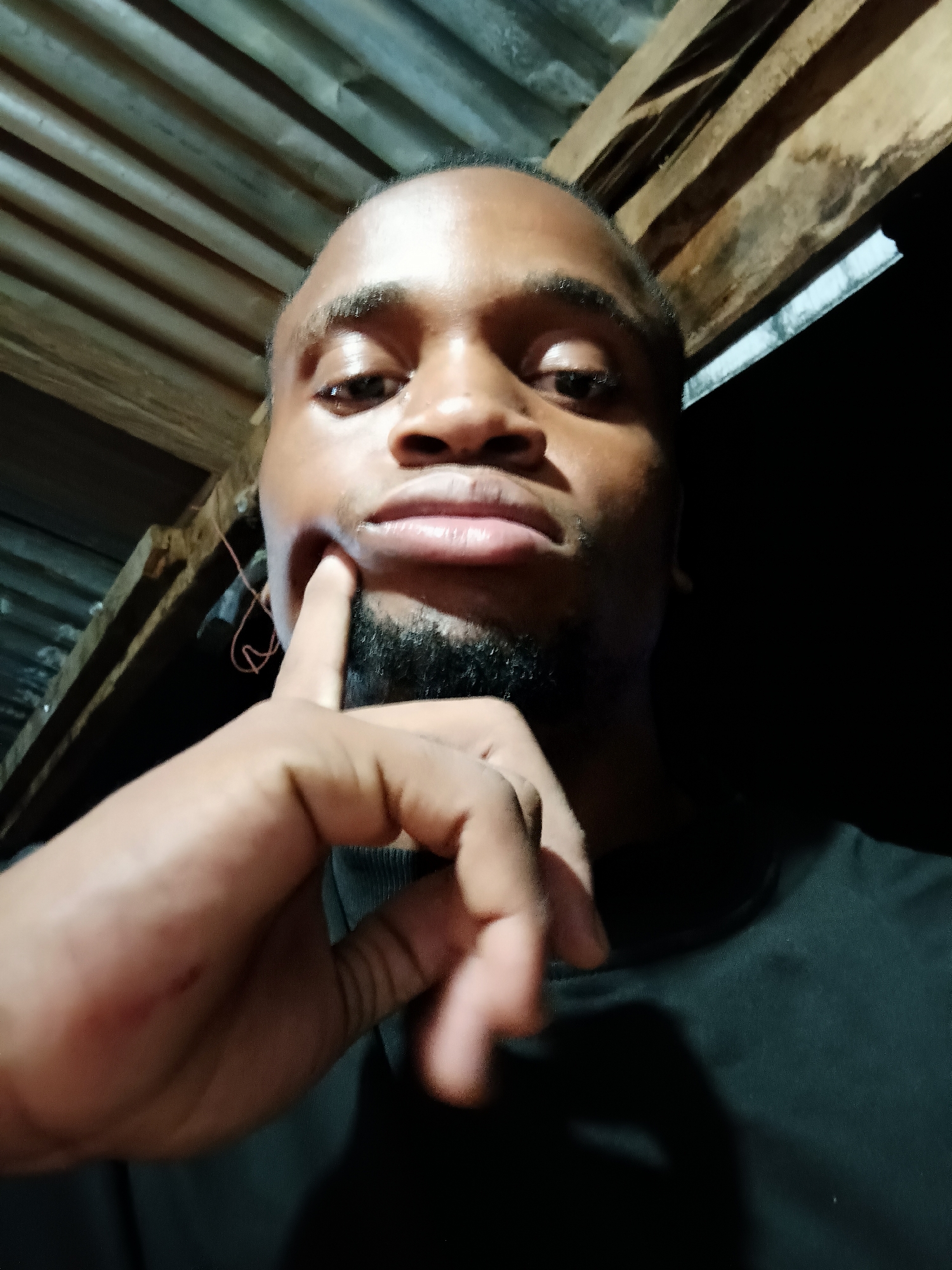 A young man with dark skin and a short beard is captured in a close-up selfie, looking directly at the camera with a neutral, possibly contemplative expression. He is wearing a black t-shirt. His left hand is raised, with his index finger touching his cheek/chin area, a pose often associated with thought or contemplation. A small scratch is visible on the palm side of his left hand, near the base of his thumb.
The setting appears to be indoors or under a simple shelter in Narayi, Nigeria, indicated by the background which features corrugated metal sheets, likely part of a ceiling or wall, and prominent wooden beams. A thin, reddish string or wire hangs near his left ear. The scene is dimly lit overall, with a strong, artificial light source directly illuminating the man's face, creating highlights on his forehead, nose, and lips, suggesting it is nighttime or a very poorly lit environment. There is no visible text in the image.