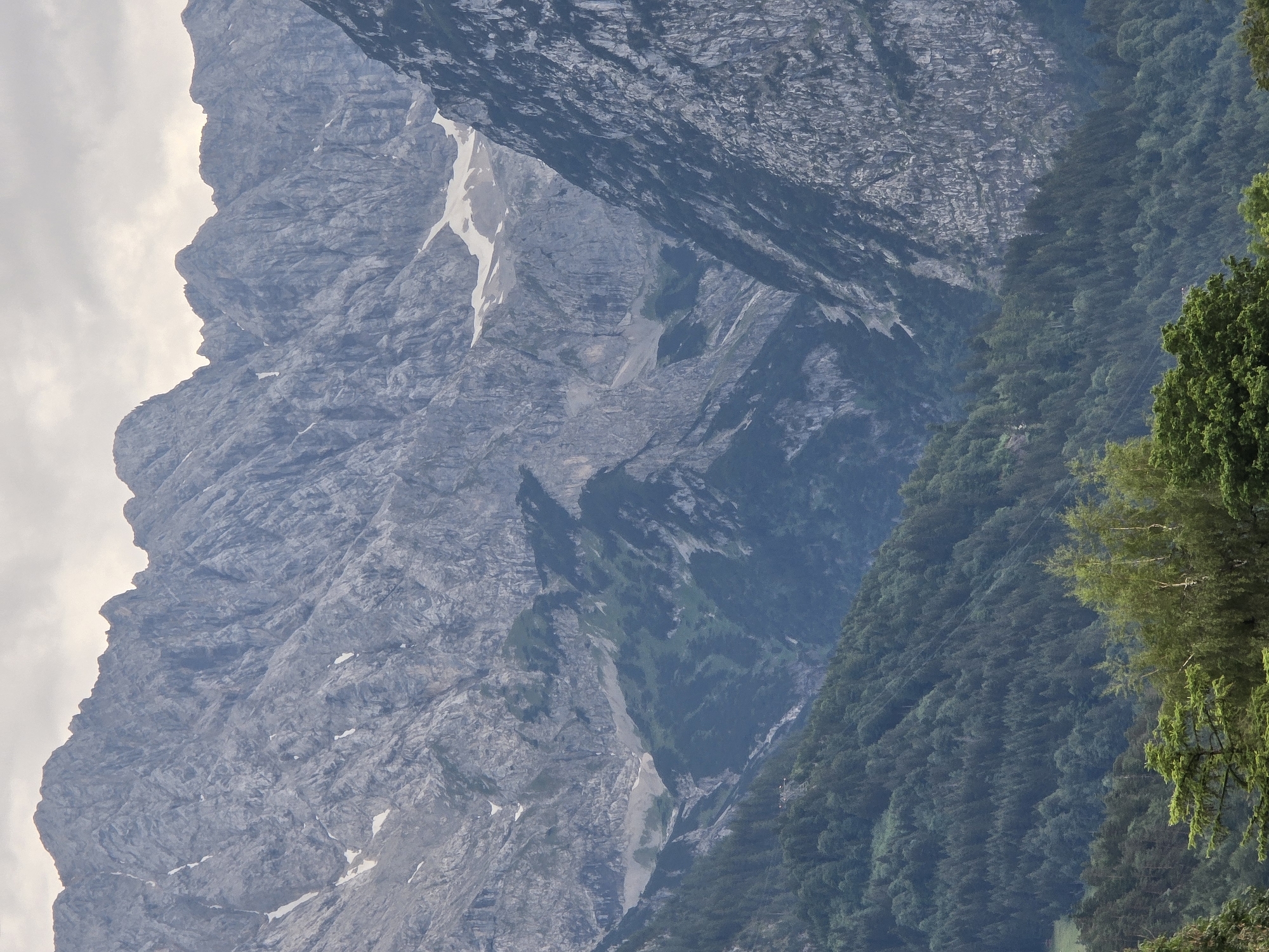 The image shows a mountainous landscape with a valley. The foreground features a dense forest of evergreen trees. In the midground, the terrain slopes upwards and is covered with more trees, with a cable car visible on the slope. The background consists of imposing, rocky mountains with patches of snow. The sky is overcast. This scene is consistent with the landscape found in Garmisch-Partenkirchen, Germany, known for its Alpine scenery.