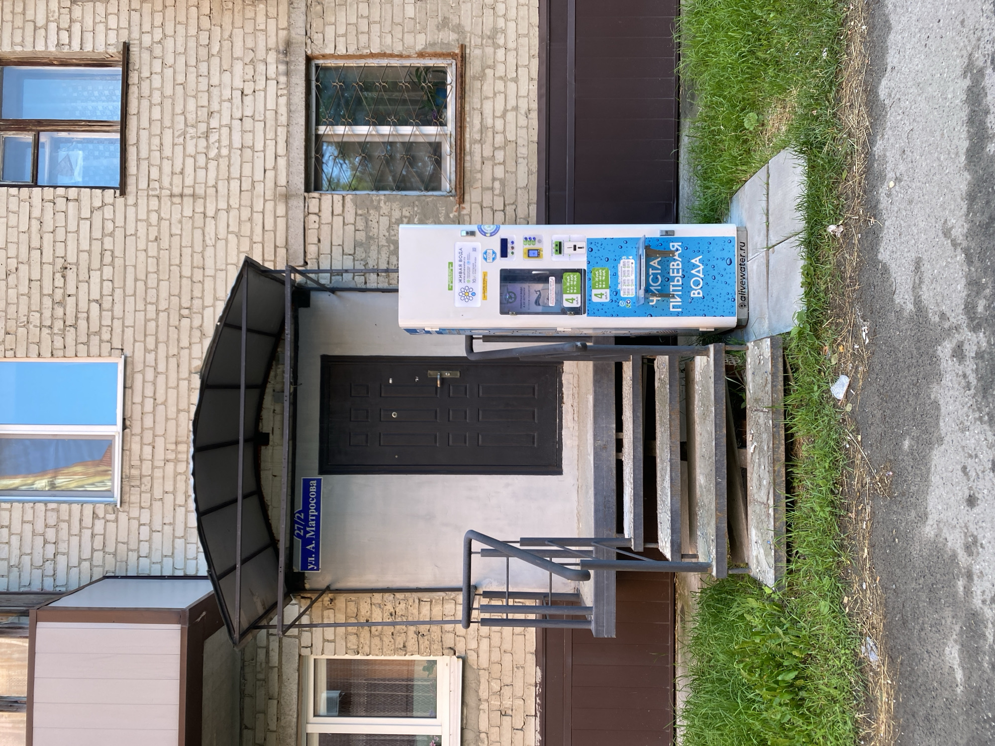 An exterior, eye-level, medium shot captures a residential building's entrance and a water vending machine. The building is constructed of light-colored bricks, with a dark brown door and a dark gray metal awning extending over a set of concrete steps leading to the door. To the left of the entrance, a window is visible, and a blue street sign indicating "ул. А. Матросова" is attached to the building's facade.
A tall, white water vending machine stands to the right of the entrance, placed on a concrete base. The machine is adorned with various stickers and logos, including one that reads "ЖИВАЯ ВОДА" (Living Water) and another with "ЧИСТАЯ ПИТЬЕВАЯ ВОДА" (Clean Drinking Water). A website address, "alivewater.ru," is also visible on the machine. The machine appears to be operational, with several buttons and a coin/bill acceptor.
The scene is set outdoors, with green grass growing along the side of the building and a paved surface, likely a sidewalk or road, to the right of the grass. The lighting suggests it is daytime, and the weather appears clear. No people are visible in the image. The overall impression is one of a functional, utilitarian setup for accessing clean drinking water in a residential area.