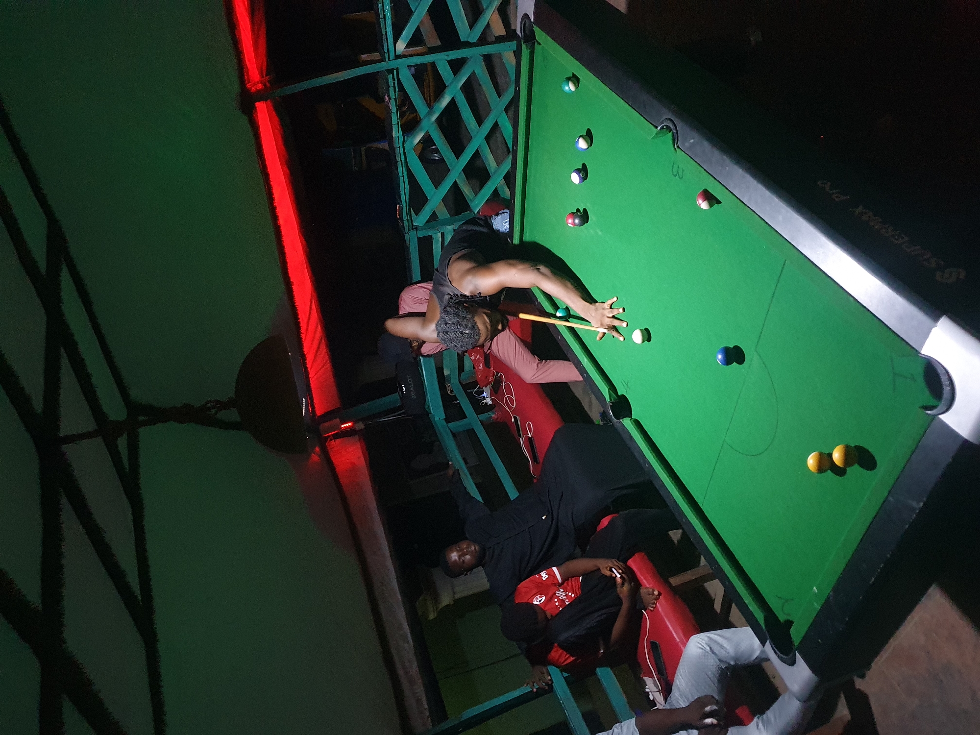 Here is a detailed description of the image:
**Location:** The image appears to be taken indoors, likely in a recreational or social space in Lokoja, Nigeria, due to the presence of individuals who appear to be Nigerian.
**People:**
* One man is actively playing pool on a green felt pool table. He is poised, holding a cue stick, and aiming at the balls.
* Several other individuals are present, observing or seated near the pool table. Some are seated on what appears to be tiered seating.
**Subjects:**
* A pool table with pool balls arranged on its surface.
* A cue stick being used by the player.
* Tiered seating structure near the pool table.
**Objects:**
* Pool balls of different colors and numbers.
* Cue stick.
* Pool table.
* Phones, possibly in use or stored.
* Lighting fixtures (overhead or spotlights).
* Tiered seating with red cushions.
* Wooden or metal railing.
**Actions:**
* A man playing pool and taking a shot.
* Other individuals watching the game.
* People sitting and interacting, possibly engaged in conversations.
**Interactions:**
* The pool player interacting with the pool table and balls.
* Possible social interactions between the observers and the player.
**Environment:**
* The environment appears to be a recreational space, possibly a bar or a game room.
* There's a green color scheme present in the environment, possibly through wall paint or other decorative elements.
* There is a mixture of natural and artificial lighting.
* There is a wooden railing and possibly tiered seating around the pool table, providing a viewing area for spectators.
