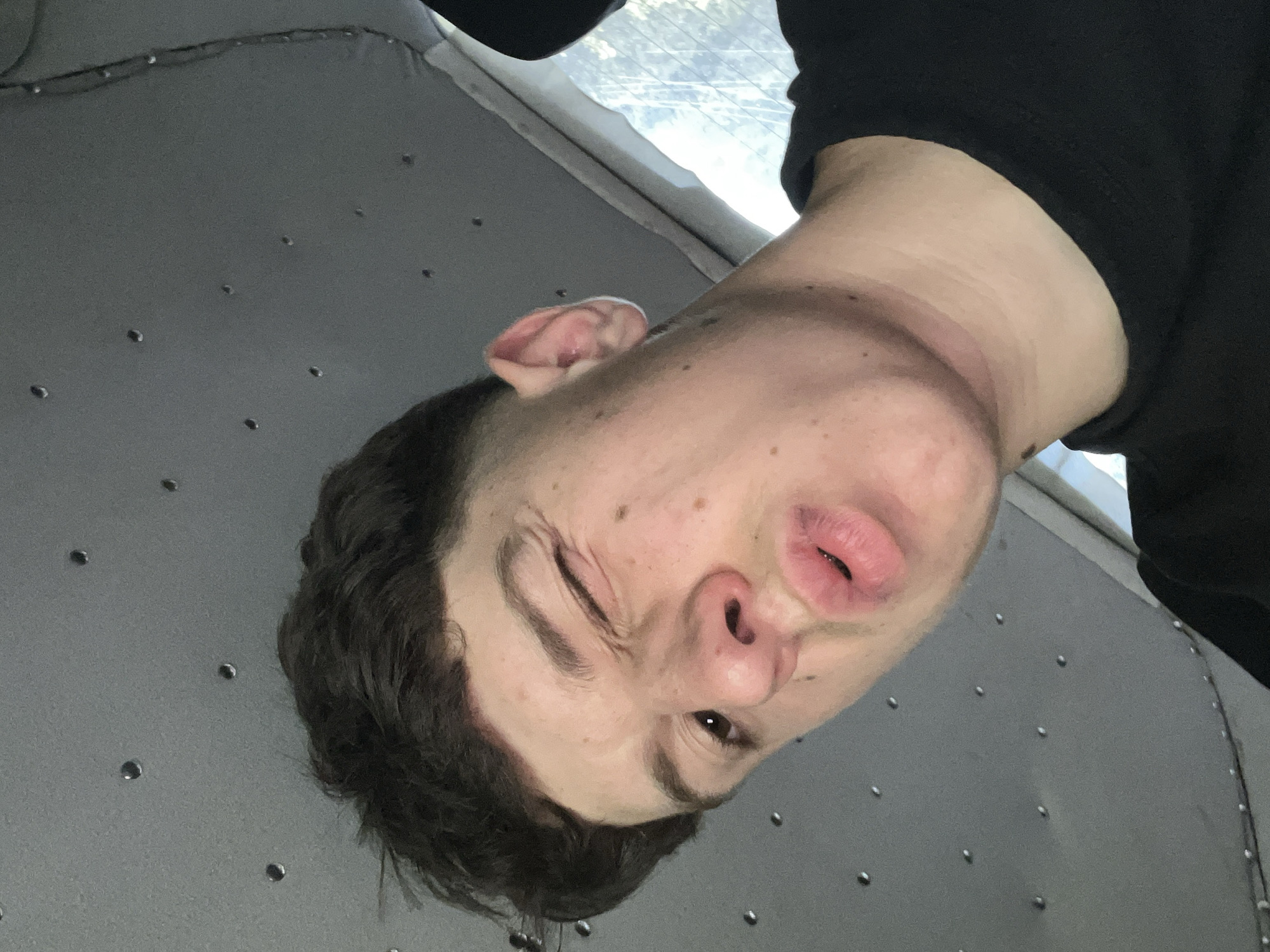A young man with dark, tousled hair is pictured from a low angle, looking towards the camera. He is winking with his left eye, and his lips are pursed. His skin appears fair with some freckles and blemishes. He is wearing a black t-shirt. The background is a grey, textured surface with circular indentations, resembling the interior of a vehicle, possibly a van or bus. A hint of a window with foliage visible can be seen to the right. The image appears to be a selfie taken indoors. There is no indication of the city of Vlahovo, Bulgaria.