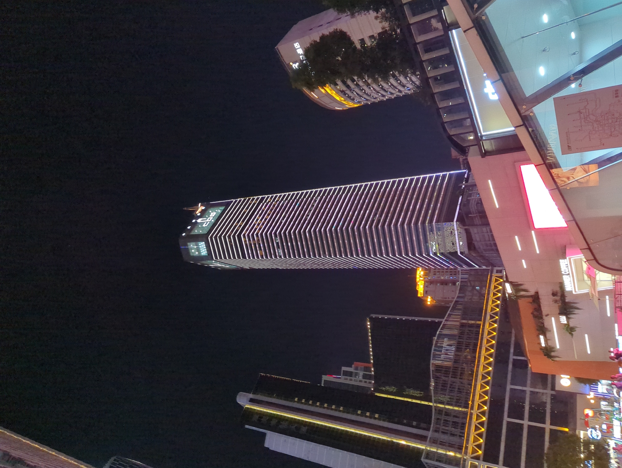 A night photograph captures a collection of modern skyscrapers and commercial buildings in an urban environment, likely in the Yuzhong District of China. The central and tallest building is a striking skyscraper illuminated with horizontal bands of white light, emphasizing its height and sleek design. To its left, another tall building displays vertical stripes of yellow light. Further to the right, a curved building is lit with orange and white lights, featuring trees on its upper levels. In the foreground, lower structures include a building with the text "MANNER COFFEE" in neon pink and a glass-enclosed entrance to a subway or underground walkway. The overall scene suggests a bustling commercial and business district at night, characterized by modern architecture and vibrant illumination.