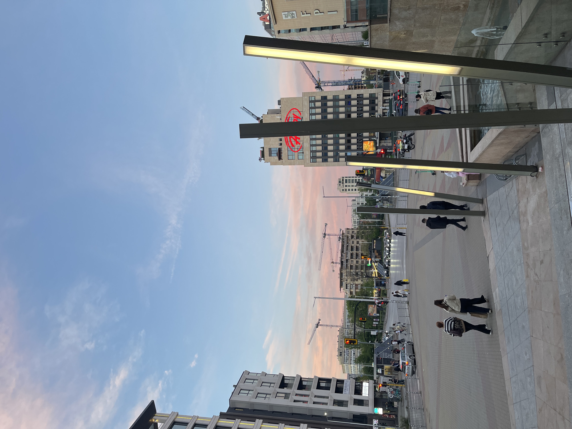 The image captures an outdoor scene in Sofia, Bulgaria, during a late day or early evening. The sky is a blend of blue and pink hues, suggesting either sunset or sunrise. Buildings of various heights and architectural styles line the street, some with construction cranes nearby, indicating ongoing development. Pedestrians can be seen walking along the sidewalk, some individually and others in groups. The illumination comes from street lights and building lights, casting shadows on the street. The image is taken from a high vantage point, looking down onto the street. A set of tall lamp posts with illuminated horizontal bars run down the right side of the image, possibly acting as a decorative feature or a divider.
