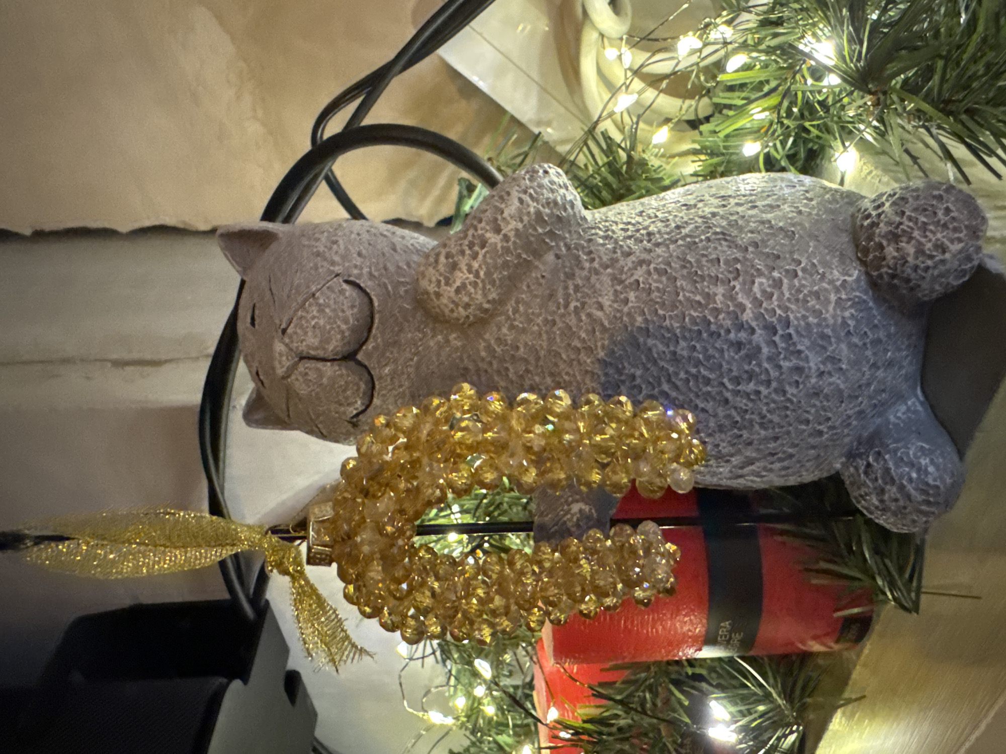 An ornamental gray cat figurine is positioned on a wooden surface, holding a golden beaded decoration in its paws. The figurine is surrounded by a string of small white lights and what appear to be pine branches. To the left of the figurine are two red cylindrical candles. The background is blurred and indistinct, with what seems to be a dark object and a wall. The image captures a festive, possibly holiday, setting. The location is not discernible from the image.