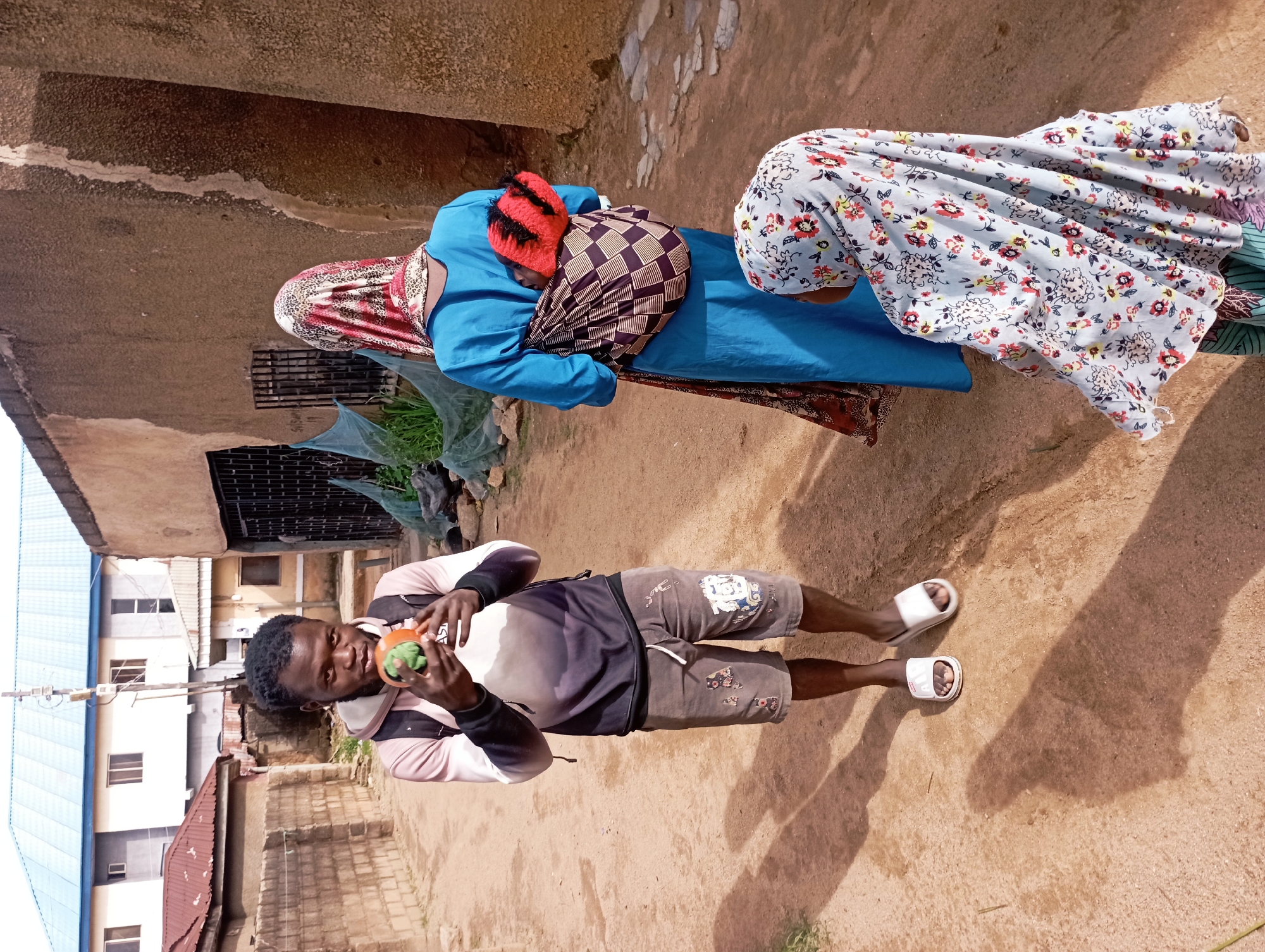 In Jos, Nigeria, a young man with dark skin and black hair holds a vibrant orange and green object, possibly a fruit or toy, in his hands, looking directly at the camera. He is wearing a gray hoodie with a white and black ombre design, gray shorts with patterned patches, and white slide sandals. To his right, a woman in a bright blue dress and a red and white patterned headscarf is seen from the back, carrying a baby wrapped in a patterned cloth on her back. The baby is wearing a red and black striped hat. A young child wearing a light blue headscarf with a floral pattern is visible in the foreground, facing away from the camera. They are standing on a dirt path in what appears to be a residential area, with buildings made of concrete and brick visible in the background. The environment is characterized by a dusty, reddish-brown ground and scattered vegetation.
