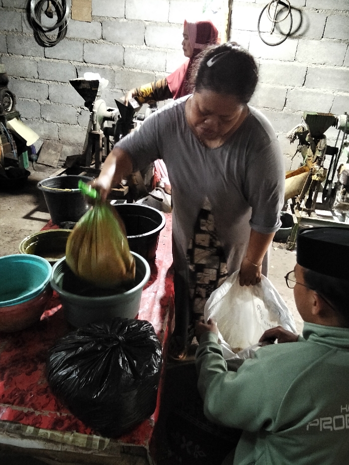 The image depicts an indoor industrial or processing area in the city of Brabe, Indonesia. Three individuals are engaged in activity within a dimly lit space with unpainted concrete block walls.
In the foreground, a middle-aged woman, wearing a grey short-sleeved top and a patterned lower garment, with her dark hair pulled back, holds a translucent green plastic bag containing a yellowish-brown, dense substance over a grey plastic tub on a red-covered table. Her left hand grasps the bag's opening, while her right hand holds the top edge of a white fabric sack presented by a man positioned to her right. This man, seen from behind, wears a black songkok and glasses, and a light green long-sleeved top with partial white lettering visible on the back. He holds the white sack open with both hands, positioned to receive contents.
To the left of the main interaction, on the red-covered table, are an aqua blue plastic bowl stacked on a red plastic bowl, and a tied-off black plastic bag. Behind the grey tub, two black plastic buckets are visible, one containing a dark, viscous material.
In the background, a second woman, wearing a red head covering and a patterned shirt, operates a piece of industrial machinery, likely a grinder or press with a hopper, mounted on a stand. Another similar machine is visible further to the right. Black electrical cables are coiled and affixed to the concrete wall in the upper left. The floor appears dark and uneven.