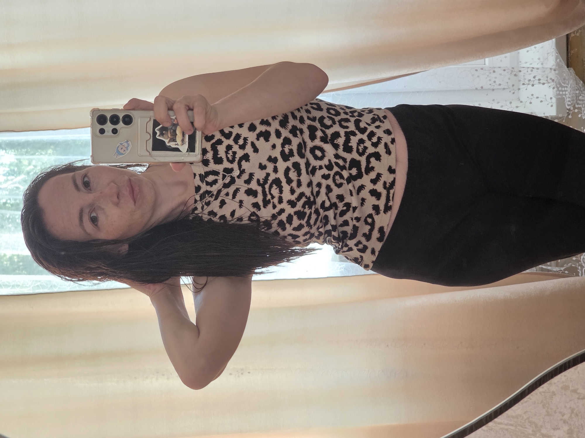 A woman is taking a selfie in a mirror. She has long, dark hair and is wearing a leopard-print tank top and black leggings. She is holding her phone up to take the picture, and her arm is bent. The background is a window with sheer curtains. The room appears to be a bedroom. The lighting is natural, coming from the window.