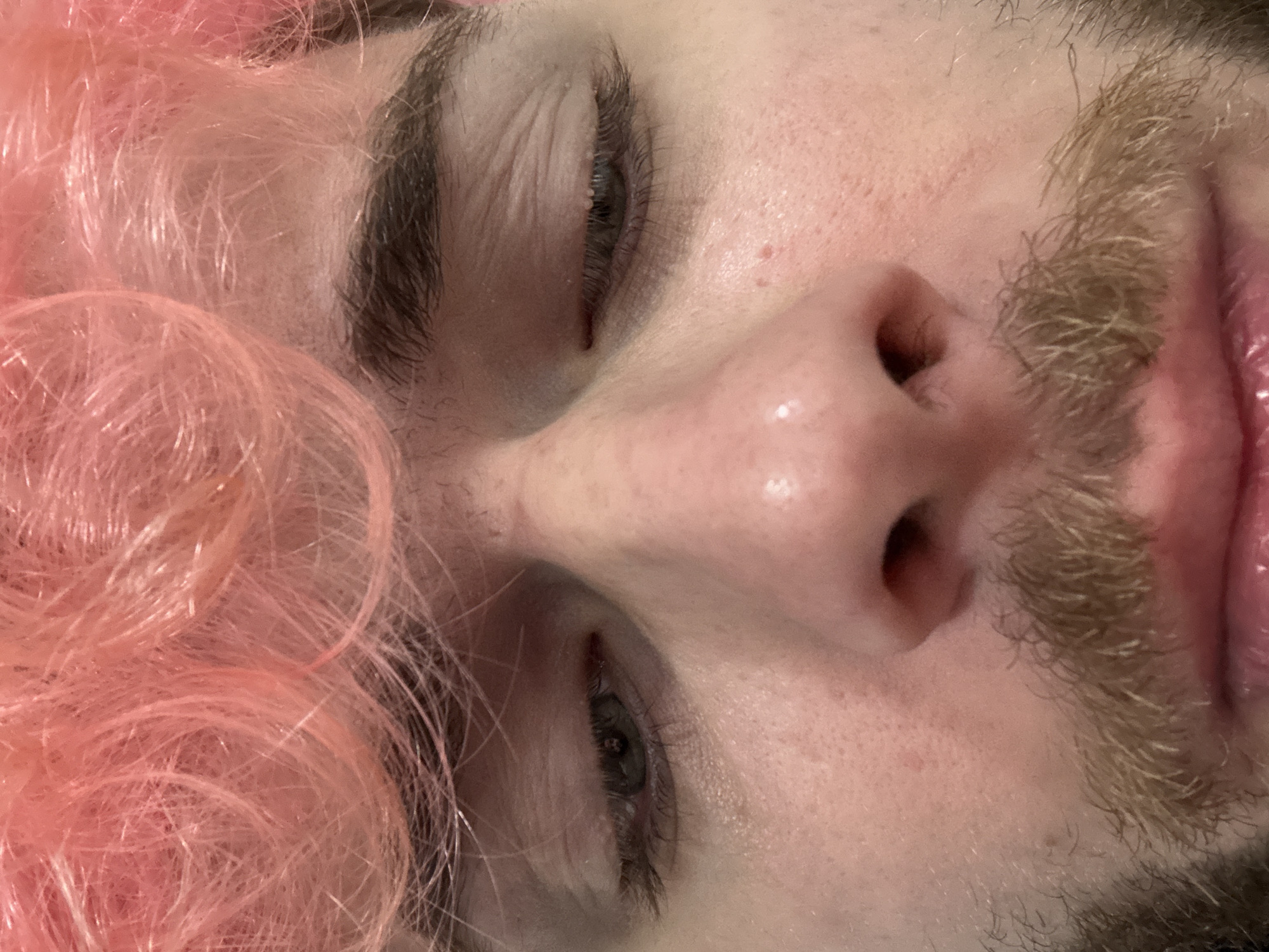 A very close-up, diagonally oriented portrait captures the central facial features of a person with light skin, vibrant pink hair, and light brown facial hair. The perspective is tight, focusing on the eyes, nose, and mouth area, with no background visible.
On the left side of the frame, masses of curly or wavy pink hair, likely a wig or dyed hair, frame the face. The hair has a bright, almost synthetic sheen. The person's eyes are partially closed or cast downwards, revealing long, dark eyelashes and dark eyebrows. The skin around the eyes and on the forehead shows fine lines and visible pores, along with a few minor blemishes.
The nose is prominent in the center, with its bridge and tip appearing slightly shiny. To the right of the nose, a light brown mustache is visible, dense with individual hairs. Below the mustache, a section of the upper lip is visible, appearing plump and glossy. Further down and to the right, some stubble or beard growth is also present.
The image is a detailed study of textures—the fine strands of pink hair, the individual hairs of the mustache and eyebrows, and the nuanced surface of the skin. The expression appears neutral or contemplative, with no strong emotions conveyed due to the partially closed eyes and tight crop. There is no visible text in the image. The provided location context "Đà Nẵng, Vietnam" is not discernible from the visual content.