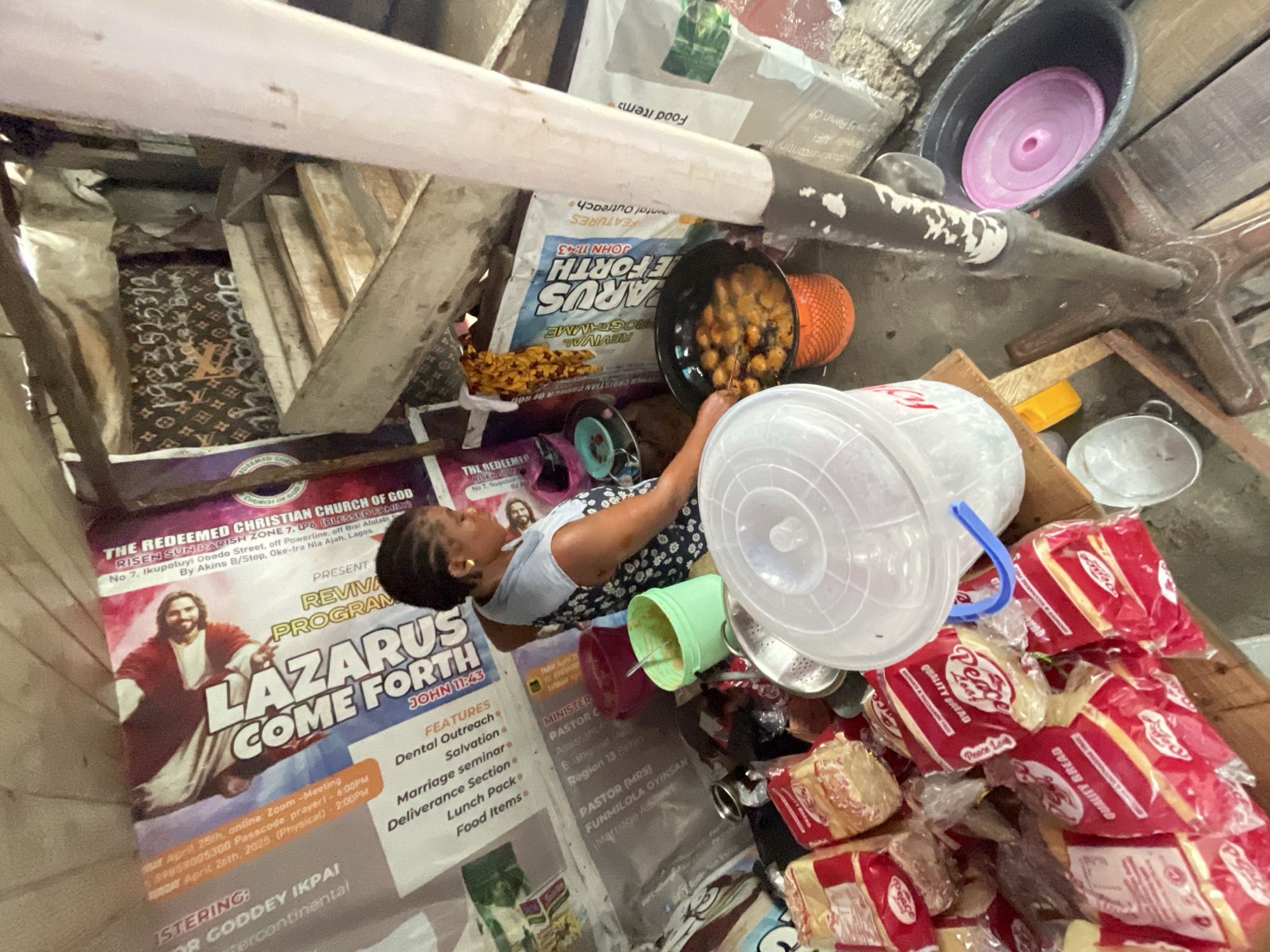 The scene appears to be a cluttered, makeshift food distribution point in Nigeria, possibly in or near the city of Ado, based on the "Oke-Ira Nia Ajah. Lagos" inscription. A woman wearing a dress is seen near a black bowl of fried foods on a Redemption Church of God flyer; The flyer advertises a "Revival Program" called "Lazarus Come Forth" with the date April 26th, 2025. The flyer also mentions salvation, dental outreach, marriage seminar, deliverance section, lunch pack, and food items. Large clear and other plastic buckets are visible, along with a stack of individually packaged loaves of "Peace" brand quality bread. Other containers, food items, and miscellaneous objects contribute to the disorganized state of the space.