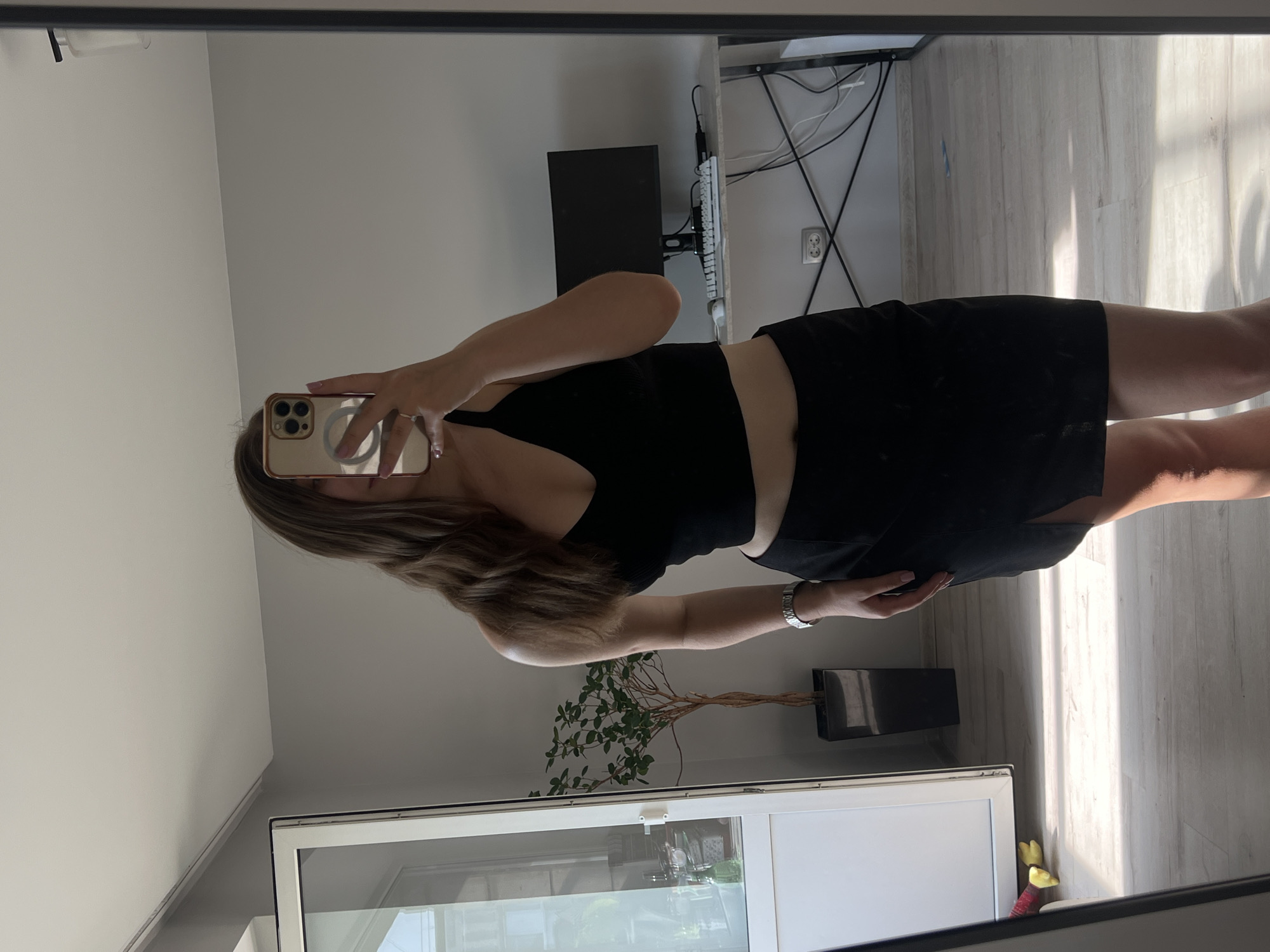 A young woman with long brown hair is taking a mirror selfie in a room. She is wearing a black crop top and a black skirt with a slit. The room appears to be a bedroom or living area, with a large mirror, a plant in a pot, a desk with a computer monitor and keyboard, and a doorway with a white door. Sunlight is streaming in from a window, casting shadows on the floor. The location is identified as Ternopil, Ukraine.