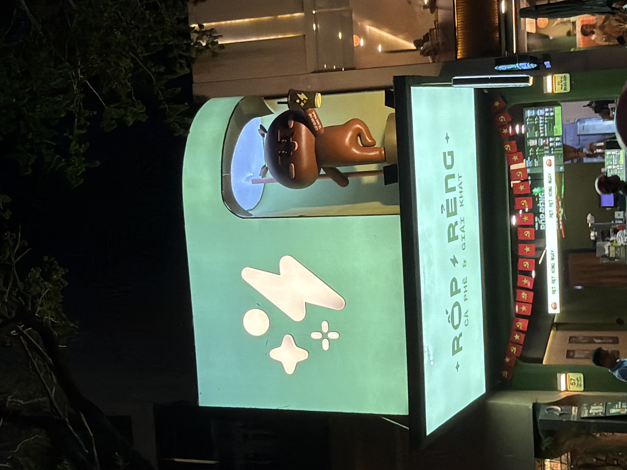 This is an outdoor nighttime shot of a quirky cafe or shop in Đà Nẵng, Vietnam. The central focus is a large, light green, somewhat futuristic-looking structure with a display window. Inside the window, a brown, cartoonish character with large eyes and a flattened nose is suspended, wearing a small brown hat with yellow details and what appears to be a yellow canister on its head.
To the right of the character's display, a vertically oriented sign features large white text that reads "+ RỐP RẾNG" and smaller text below it "CÀ PHÊ GIẢI KHÁT," indicating it's a coffee and beverage shop. Along the right edge of this green structure, a string of small red flags with yellow stars and communist symbols is draped. Above these flags, there's another sign with Vietnamese text, and further to the right, a display with what looks like a menu or products is visible, with people blurred in the background.
On the left side of the green structure, there are white graphic elements resembling abstract shapes like a lightning bolt, a circle, and a starburst, all against the light green background. In the foreground on the left, dark tree branches with green leaves are visible, suggesting the cafe is located amongst some greenery, possibly in a courtyard or outdoor seating area. The overall mood is vibrant and slightly playful, typical of a modern urban establishment in Vietnam.