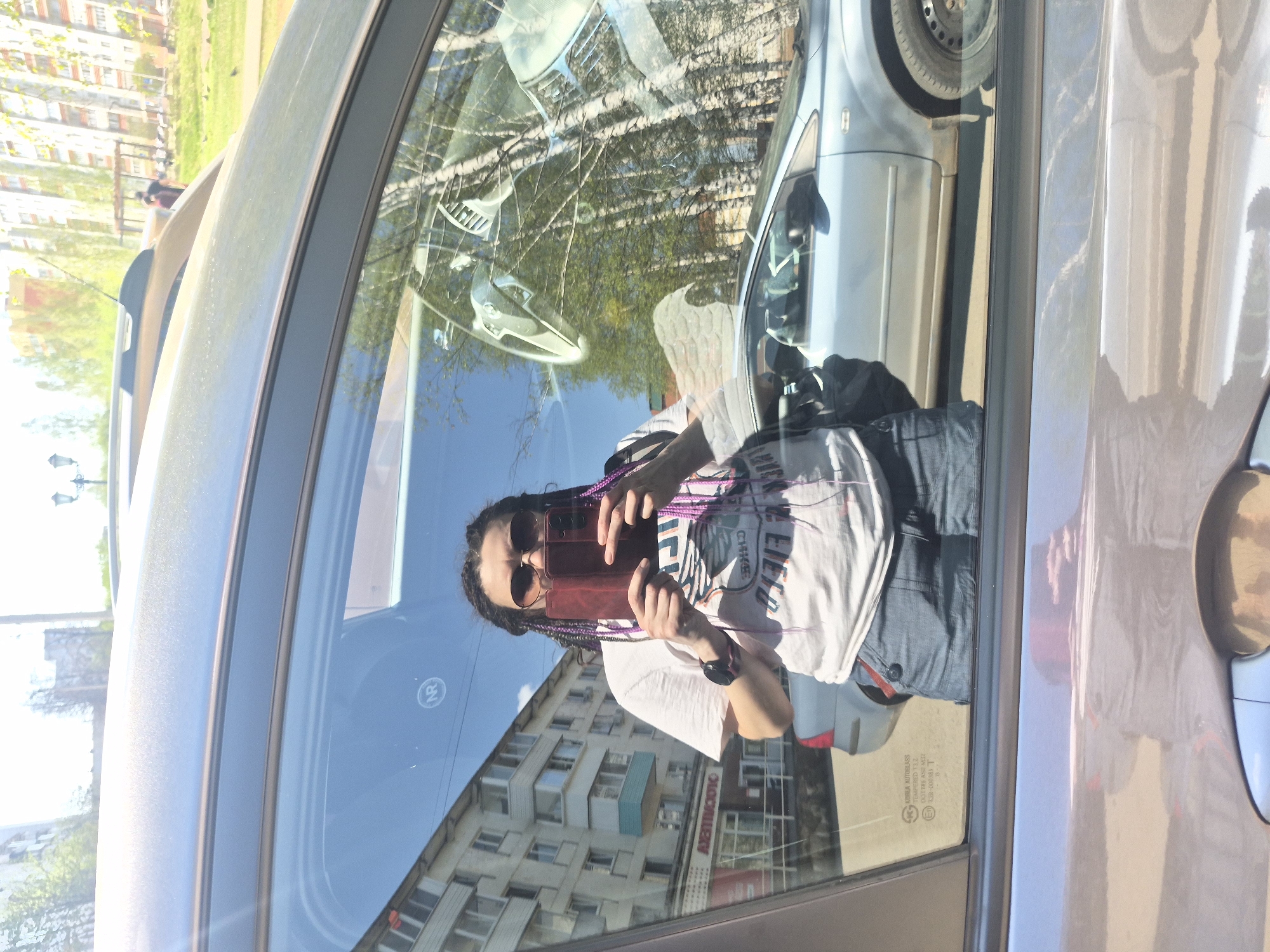 The image shows a person taking a photo of themselves in a car window. The person has dark hair in braids, is wearing sunglasses, a white t-shirt, and dark blue jeans. They are holding a dark red phone. The reflection in the car window shows a building with multiple windows. The building has a storefront on the ground level with a sign in Russian. In the background, a park with trees and a walking path are visible. A parked silver car is also reflected in the window. The scene appears to be located in a city, likely in an urban area.