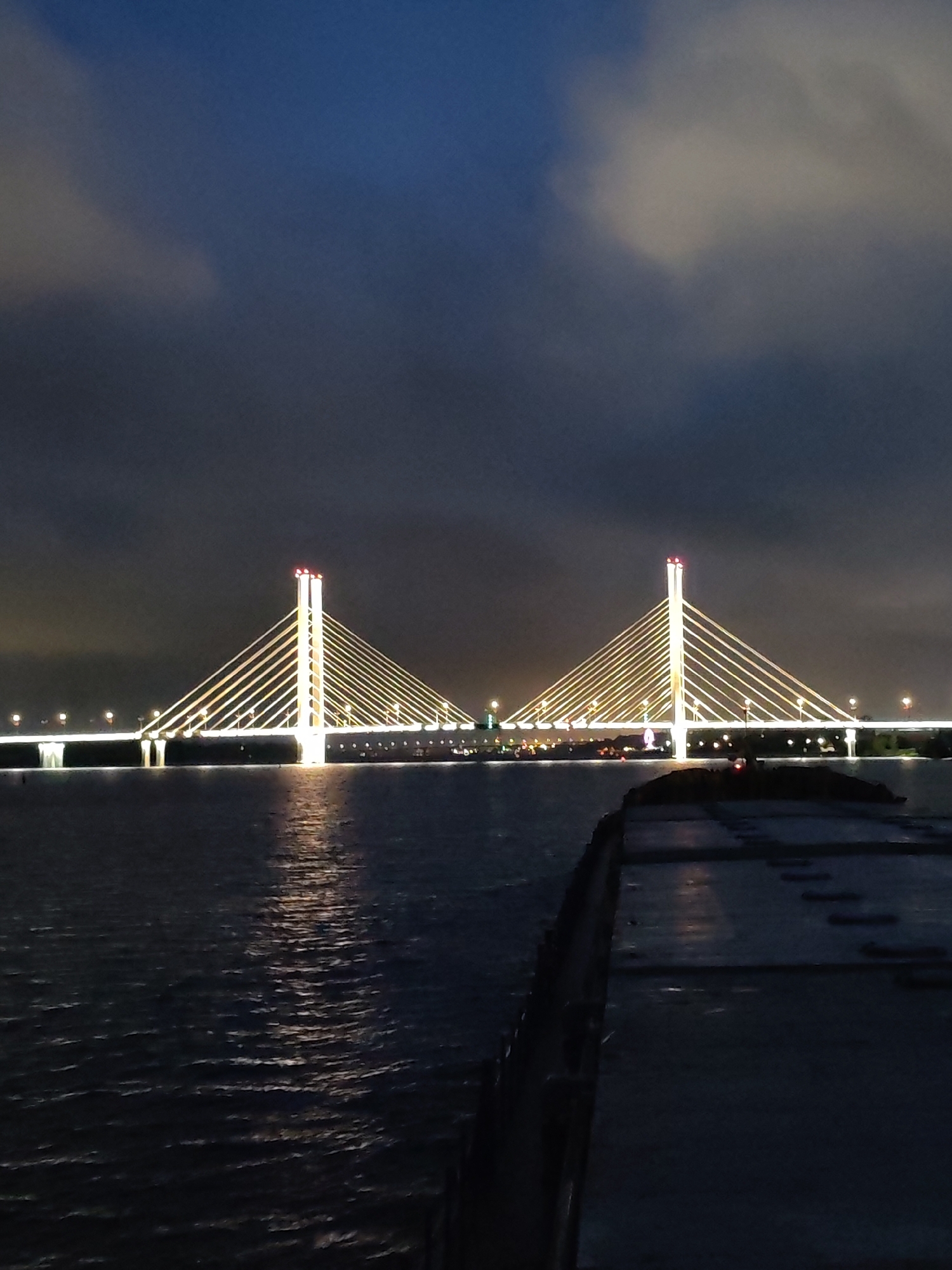 The image shows a cable-stayed bridge illuminated at night. The bridge has two towers with bright lights and cables stretching down to the roadway. Lights line the deck of the bridge. The bridge is over water with reflections of the lights visible. In the foreground, there is a dock or platform surface. Above, the sky is dark with some cloud cover. This is most likely the October Bridge in Cherepovets, Russia.