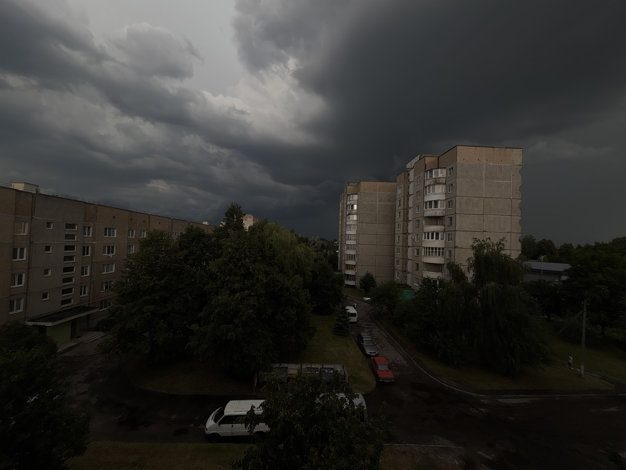 This media file captures a dramatic, overcast scene over a residential area in Lida, Belarus. The sky is filled with dark, heavy storm clouds, suggesting an imminent or ongoing weather event, creating a somber and intense atmosphere.
The foreground and midground are dominated by apartment buildings, typical of Eastern European urban landscapes. One building on the left is a light-colored, multi-story structure with many windows. Another on the right is a taller, grey concrete building, also featuring numerous windows and balconies, some of which reflect the dim light. Lush green trees are interspersed between the buildings and surround the scene, their foliage appearing dark and saturated under the oppressive sky.
A street winds through the complex, wet and reflecting the dark sky, indicating recent or ongoing rain. Several cars are parked along the street, including a white van in the lower left, a red sedan, and a darker grey sedan. The overall setting appears to be a courtyard or residential street within a housing complex.
The time of day is difficult to pinpoint precisely due to the cloud cover, but the low light suggests it could be late afternoon or early evening, coinciding with the approaching storm. There are no visible people, but the presence of parked cars and residential buildings implies human habitation. The overwhelming impression is one of a brewing storm, with the dark clouds and wet ground contributing to a mood of anticipation or unease. No text is visible in the image.