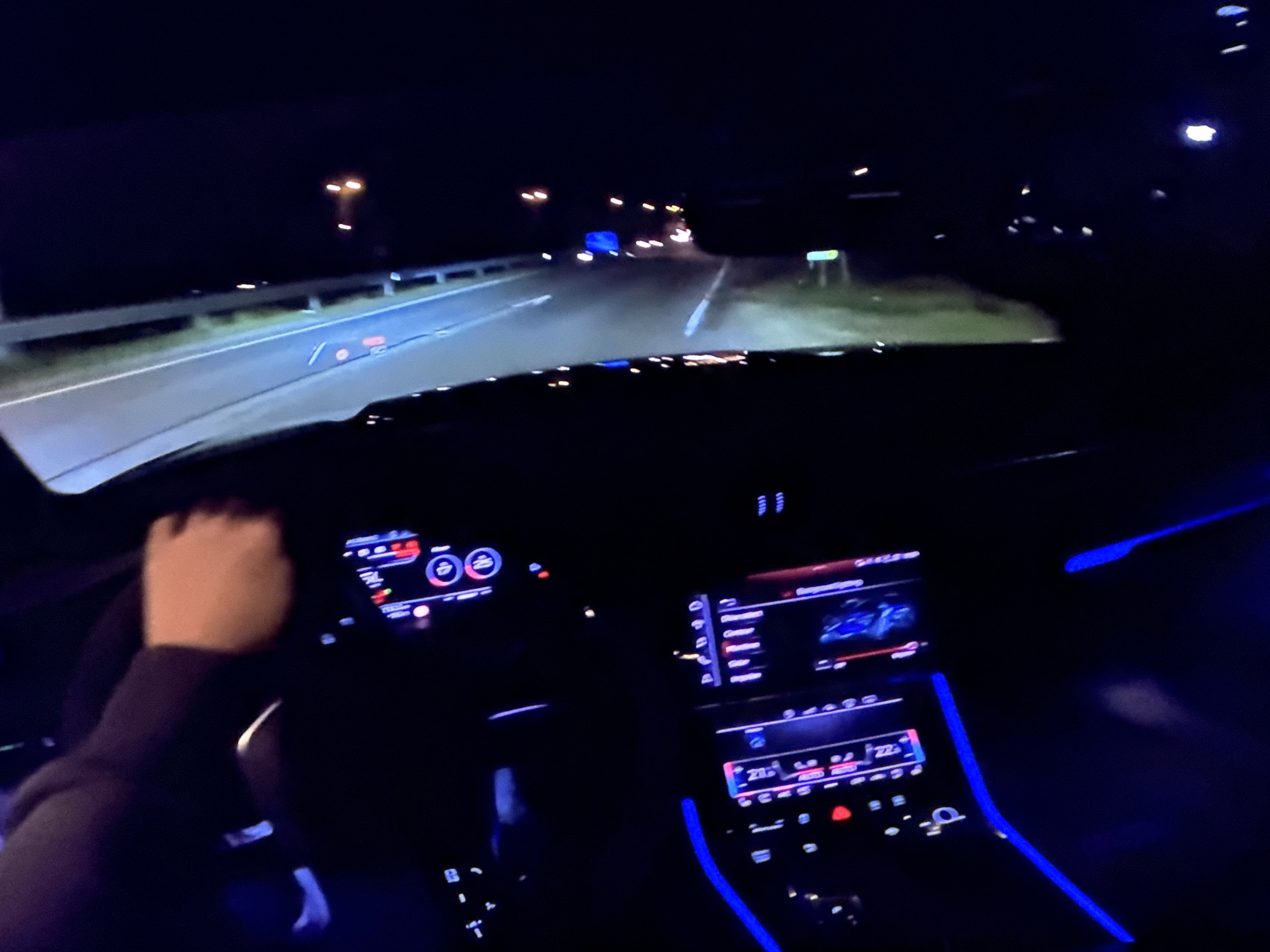 The image captures a first-person perspective from inside a modern car at night, likely on a highway. The dashboard displays are illuminated with blue and red lighting, indicating the car is operational. A hand is visible on the steering wheel, suggesting the driver is actively controlling the vehicle. The road ahead is dimly lit by headlights and streetlights, with the typical signs and guardrails of a highway visible. The car's interior ambient lighting is a prominent feature, casting a blue glow. The scene does not provide specific visual cues to confirm the location as Bezden, Bulgaria, but it represents a common scenario of nighttime driving.