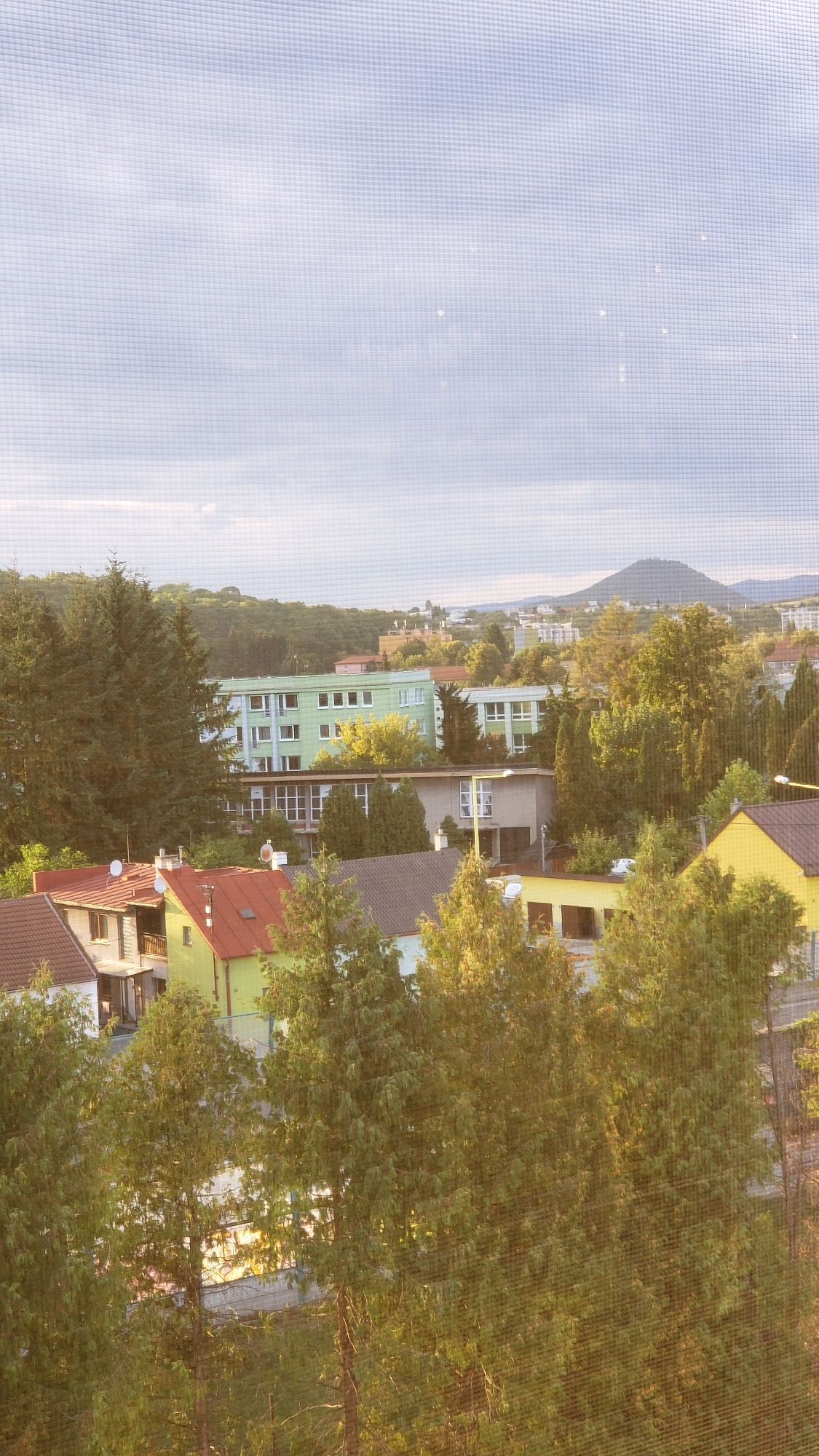 The image captures a suburban landscape in Prešov, Slovakia, likely taken from an elevated position such as a window, as evidenced by the visible screen mesh. The scene is dominated by a variety of buildings and lush greenery. In the foreground, several trees with dense foliage are visible, catching the warm light of what appears to be late afternoon or early morning.
Behind the trees, a row of houses with red and green roofs can be seen, suggesting a residential area. Further back, larger, light green buildings with multiple windows indicate a more institutional or public complex, possibly a school or dormitory. The architecture suggests a mixture of residential and potentially communal structures.
The middle ground features more trees interspersed with buildings, and a street or pathway is faintly visible. The background is characterized by a gently rolling, forested hill, and beyond that, the silhouette of a mountain or significant elevation rises against a pale, cloudy sky. The overall impression is one of a peaceful, somewhat developed area integrated with nature. The quality of light and the presence of the screen mesh imply that this is a snapshot taken from within a building, looking out at the city.
