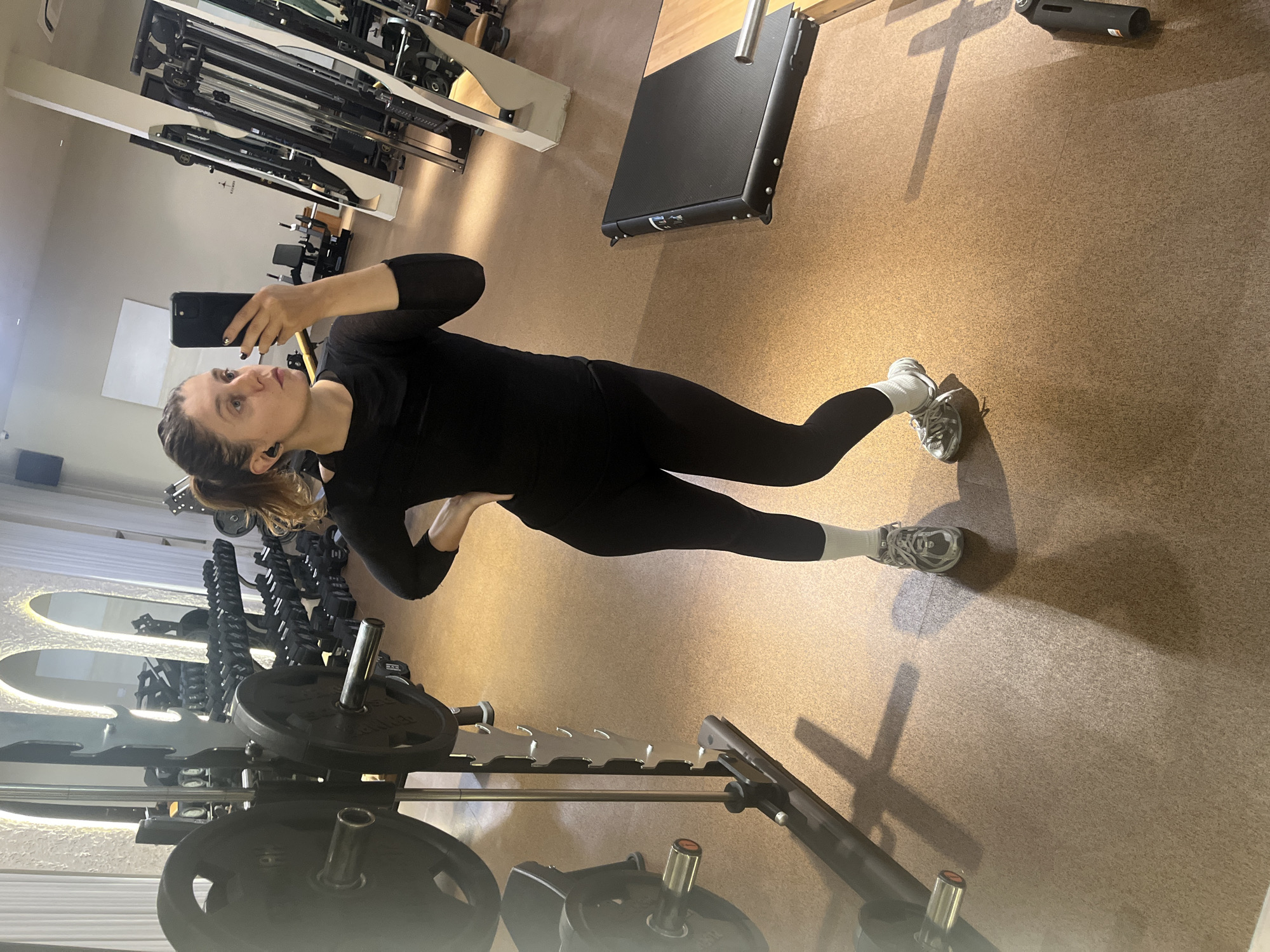 A woman in black athletic wear is taking a selfie with a black smartphone in a gym. She is wearing grey sneakers with white socks. The gym features equipment like a smith machine loaded with weight plates, a cable machine, and a platform. The flooring is a speckled brown material. The gym's location is North Kuta, Indonesia.