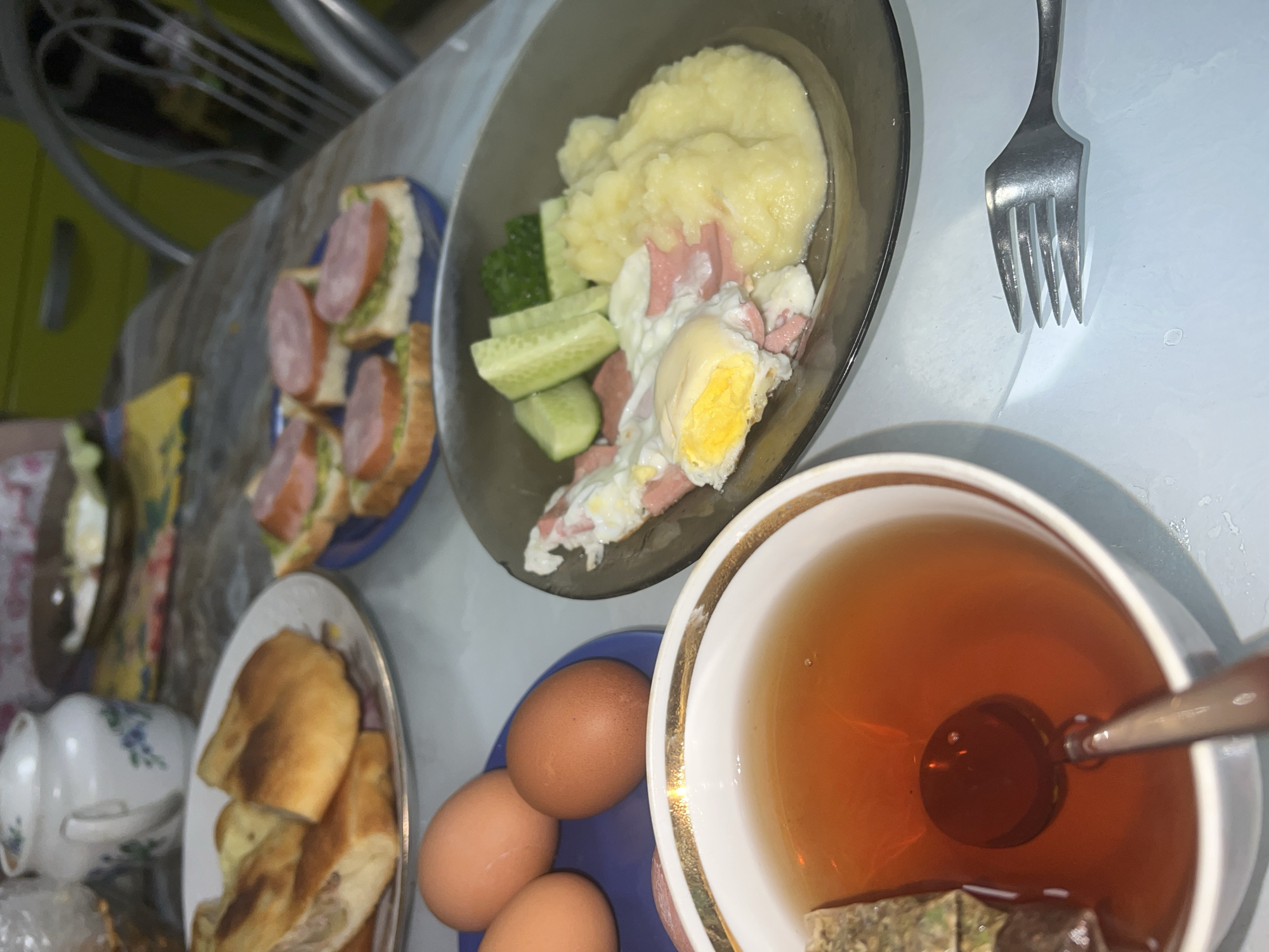The image displays a substantial meal laid out on a light-colored table, suggesting an indoor setting like a kitchen or dining area. The main dish, presented on a dark, transparent plate, consists of a generous portion of mashed potatoes, alongside several slices of fresh cucumber and a fried egg with a solid yellow yolk, accompanied by strips of cooked ham or sausage.
To the left, a blue plate holds several small, open-faced sandwiches made with white bread, a green spread, and round slices of sausage or salami. In the lower left, a white plate contains multiple golden-brown pastries, possibly fried or baked, which appear to be filled. Adjacent to these, on a smaller blue plate, are three raw brown eggs.
A white teacup with a distinctive gold rim, filled with amber-colored tea, a metal spoon, and a tea bag, is prominent in the lower right. A silver four-pronged fork rests on the table near the main plate. Partially visible in the background are a white teapot or sugar bowl adorned with a blue floral pattern, a light green surface (likely a cabinet), and a metal rack or kitchen utensil.
The diverse array of items suggests a hearty breakfast or brunch. The composition of the meal, featuring staples like eggs, potatoes, sausage, and tea, is consistent with a home-cooked meal in a domestic environment, fitting the provided context of Perm, Russia. No people or visible text are present in the image.