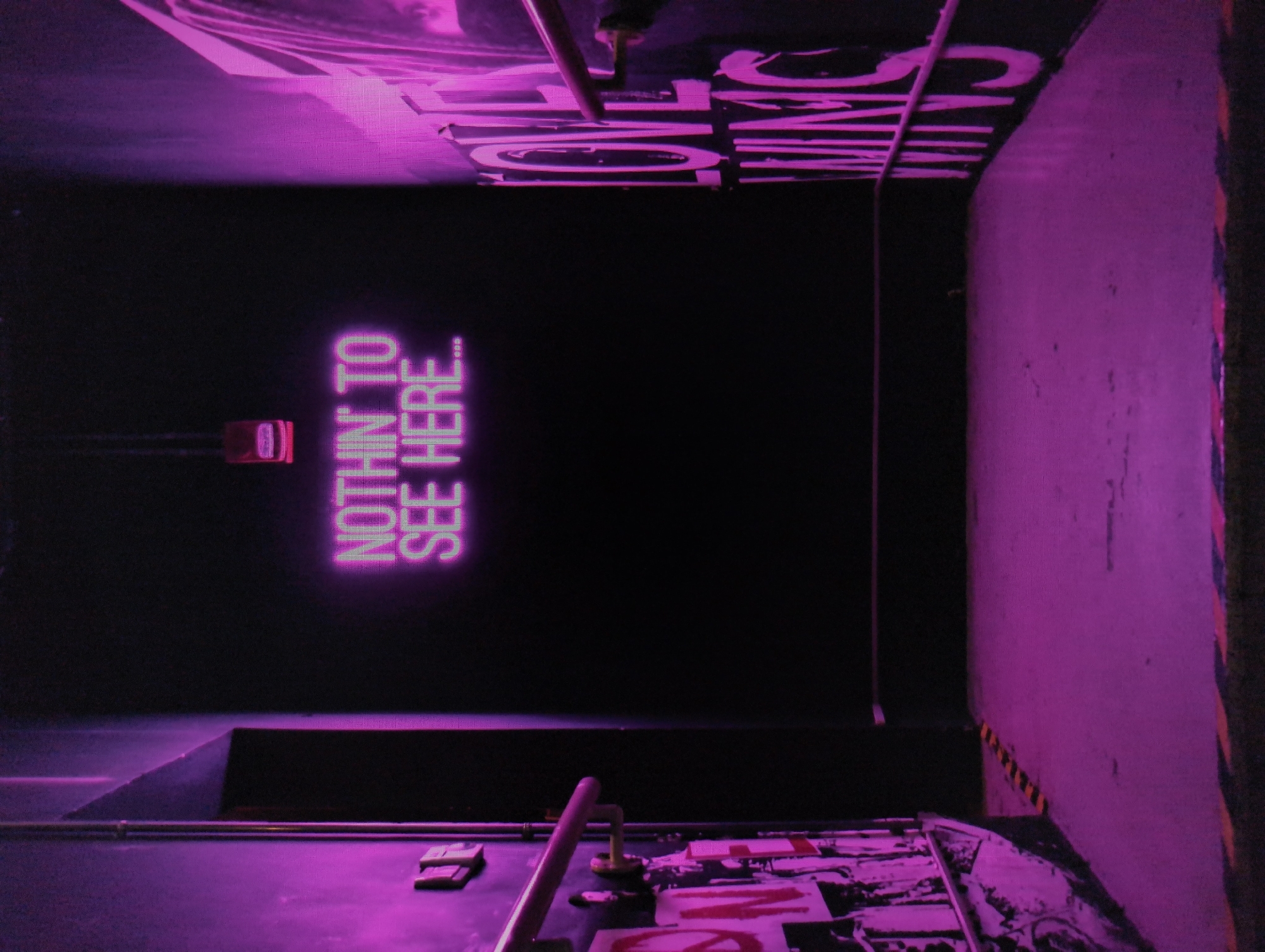 The image shows a dimly lit corridor in what appears to be an industrial or commercial space. The walls and floor are primarily illuminated in purple. A sign reading "NOTHIN TO SEE HERE..." is prominently displayed on the back wall. Safety stripes are also visible along the edge of the wall. The lighting and composition of the image suggest a clandestine or hidden area. It is not possible to specify the location or presence of people/objects/interactions from this image.