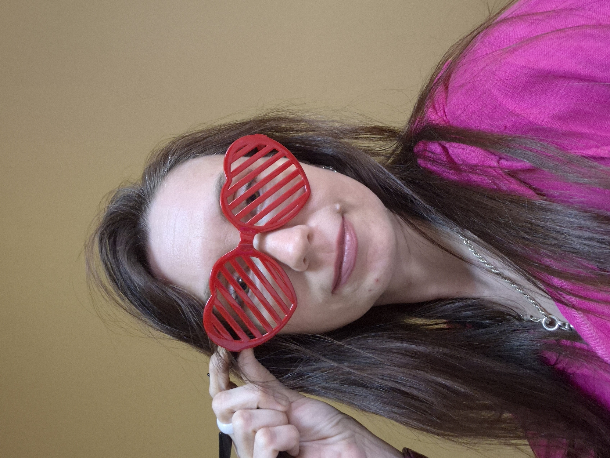 A woman with long brown hair is wearing red heart-shaped sunglasses. She is wearing a pink top and a silver necklace. She is holding the sunglasses with her left hand. The background is a plain beige color. The image appears to have been taken indoors. The location is unknown.