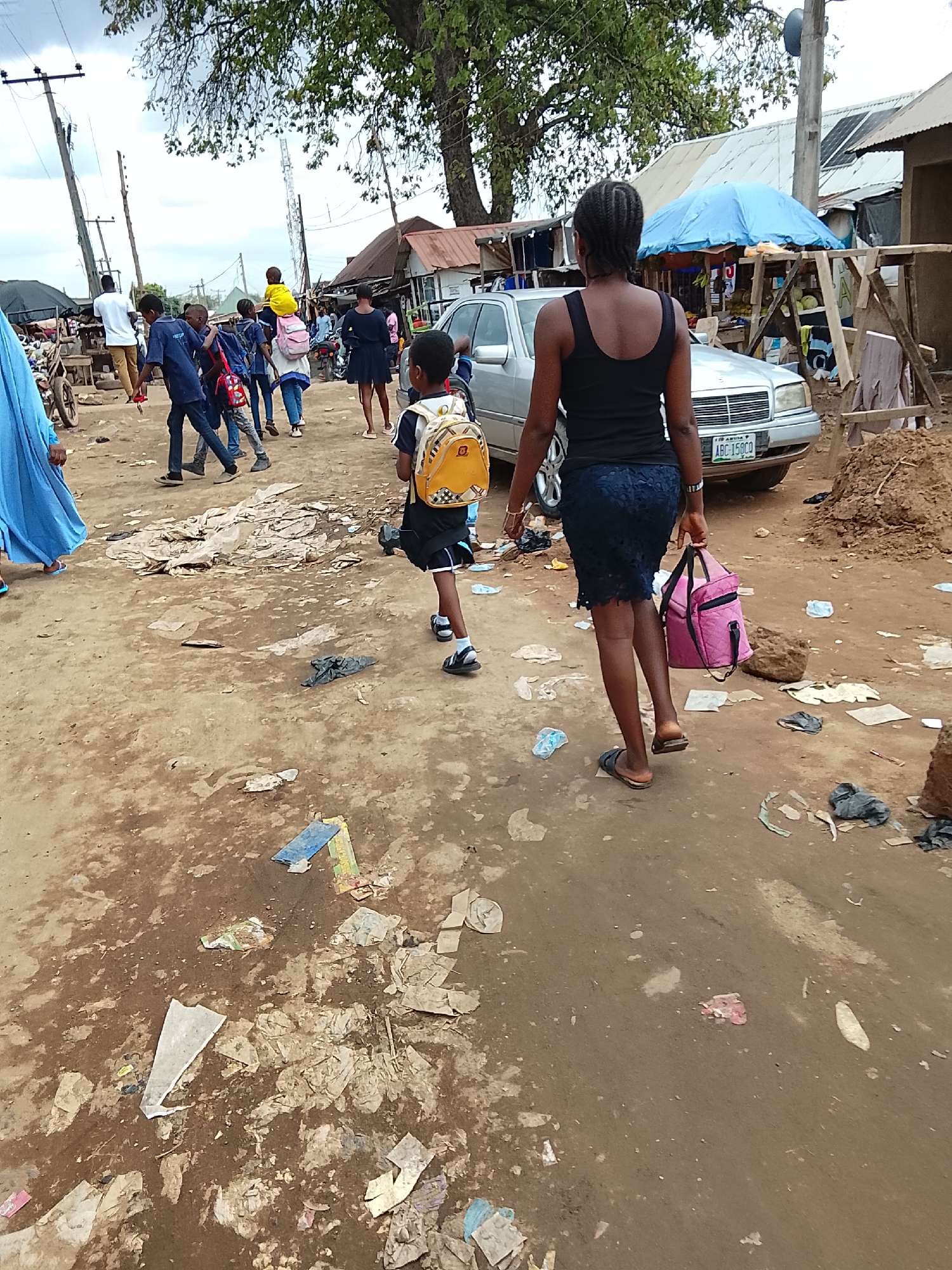The image shows a dirt road scene in what appears to be a marketplace or residential area. In the foreground, a woman and a young boy are walking away from the viewer. The woman is carrying a pink bag and the boy is wearing a yellow backpack. Several other people are walking in the same direction. A silver car is parked to the right. The ground is covered in litter and debris. Buildings are visible on both sides of the road. The scene is likely located in Abuja, Nigeria based on the car's license plate.