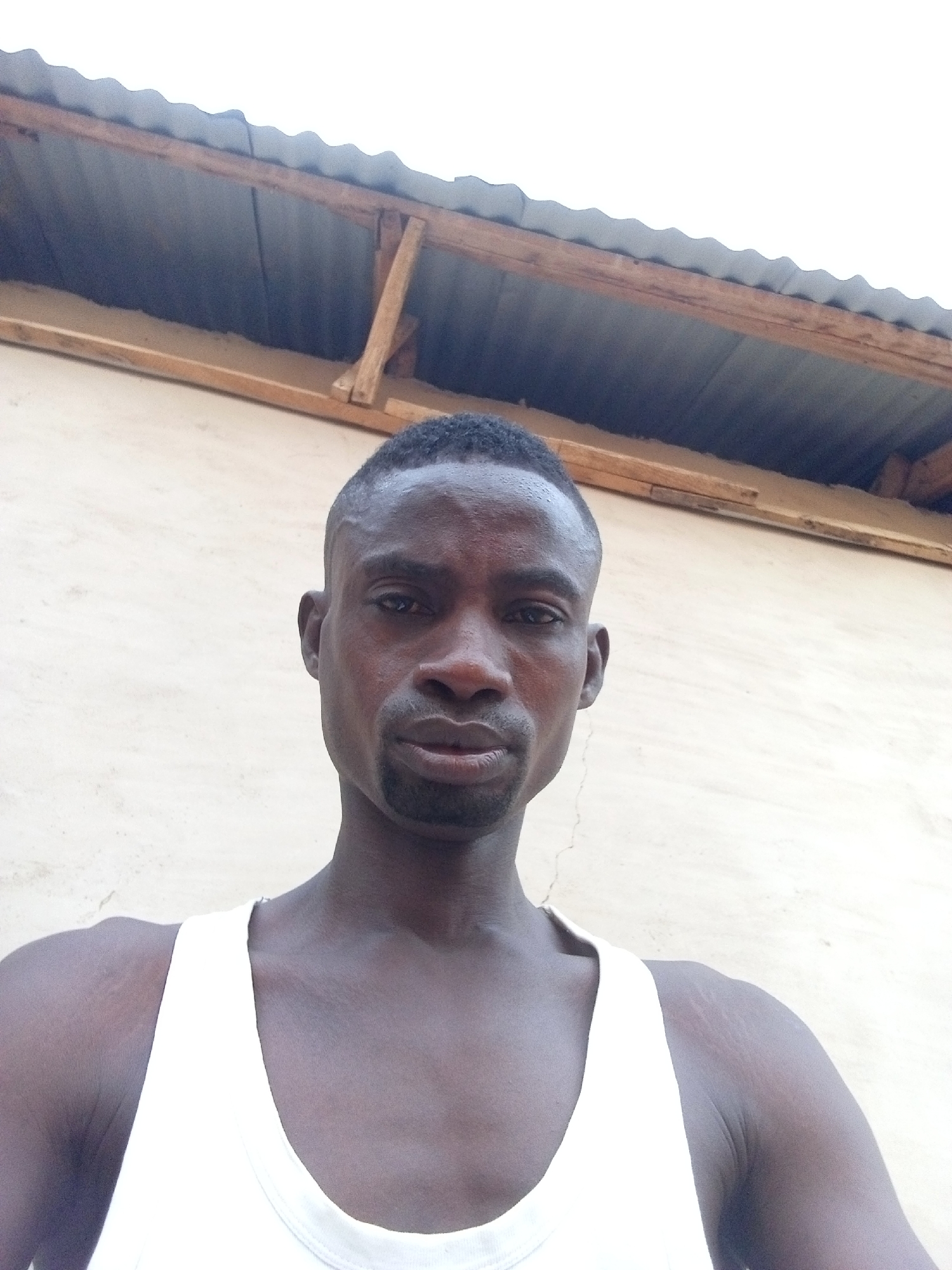 A medium shot depicts a dark-skinned man wearing a white tank top. He has a short, closely cropped haircut and a goatee. The background features a beige wall and a corrugated metal roof structure with wooden supports. The location is identified as Unknown, Unknown.