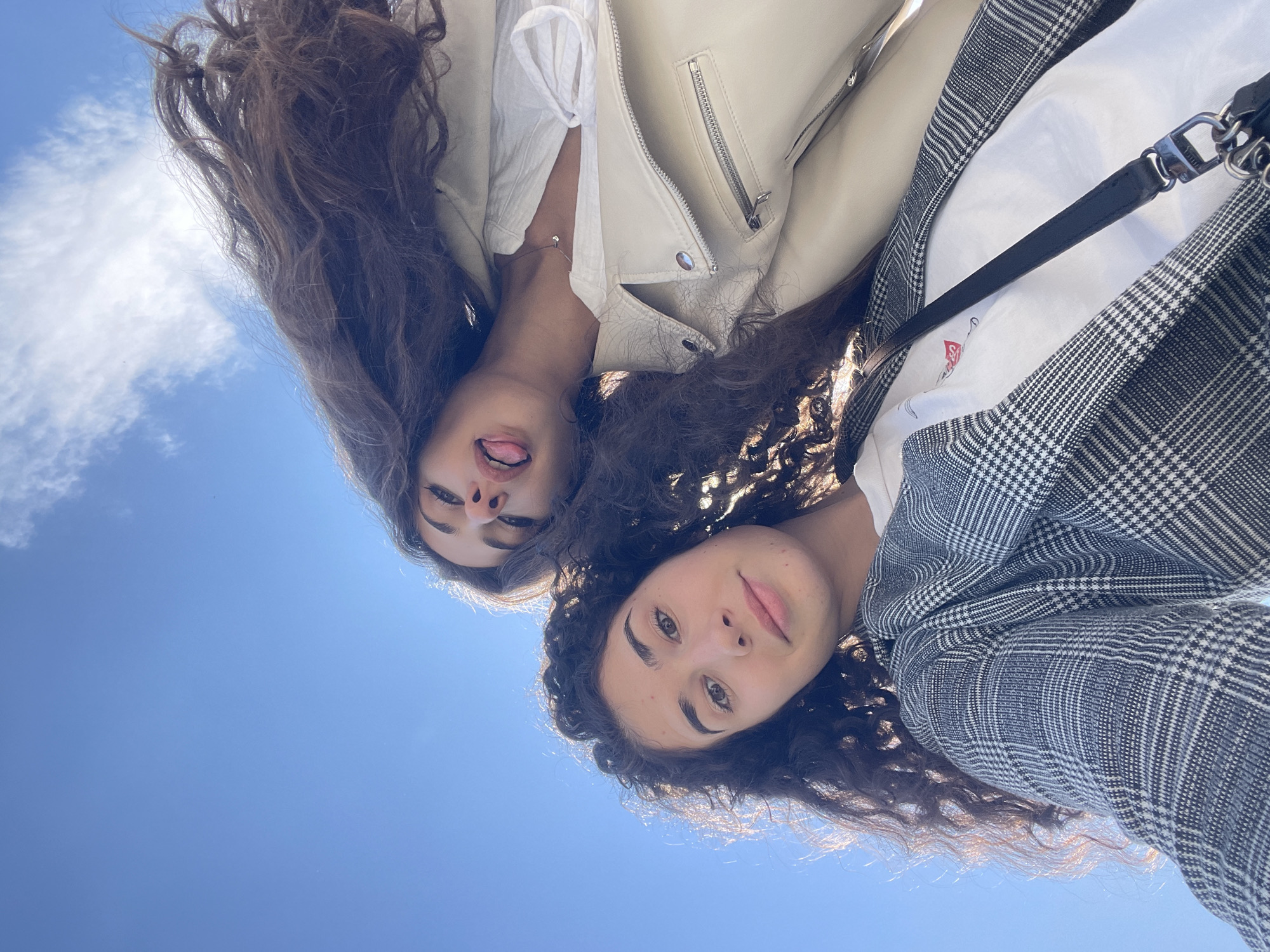 Two young women are pictured against a clear blue sky with scattered clouds. The camera is angled upwards, giving a worm's-eye view. The woman on the right wears a grey plaid jacket over a white shirt. The woman on the left wears a cream-colored leather jacket over a white top. Her tongue is visible. The scene suggests an outdoor location, based on the open sky, but does not definitively indicate a location in Sofia, Bulgaria or provide any forensic detail on the interaction between them.