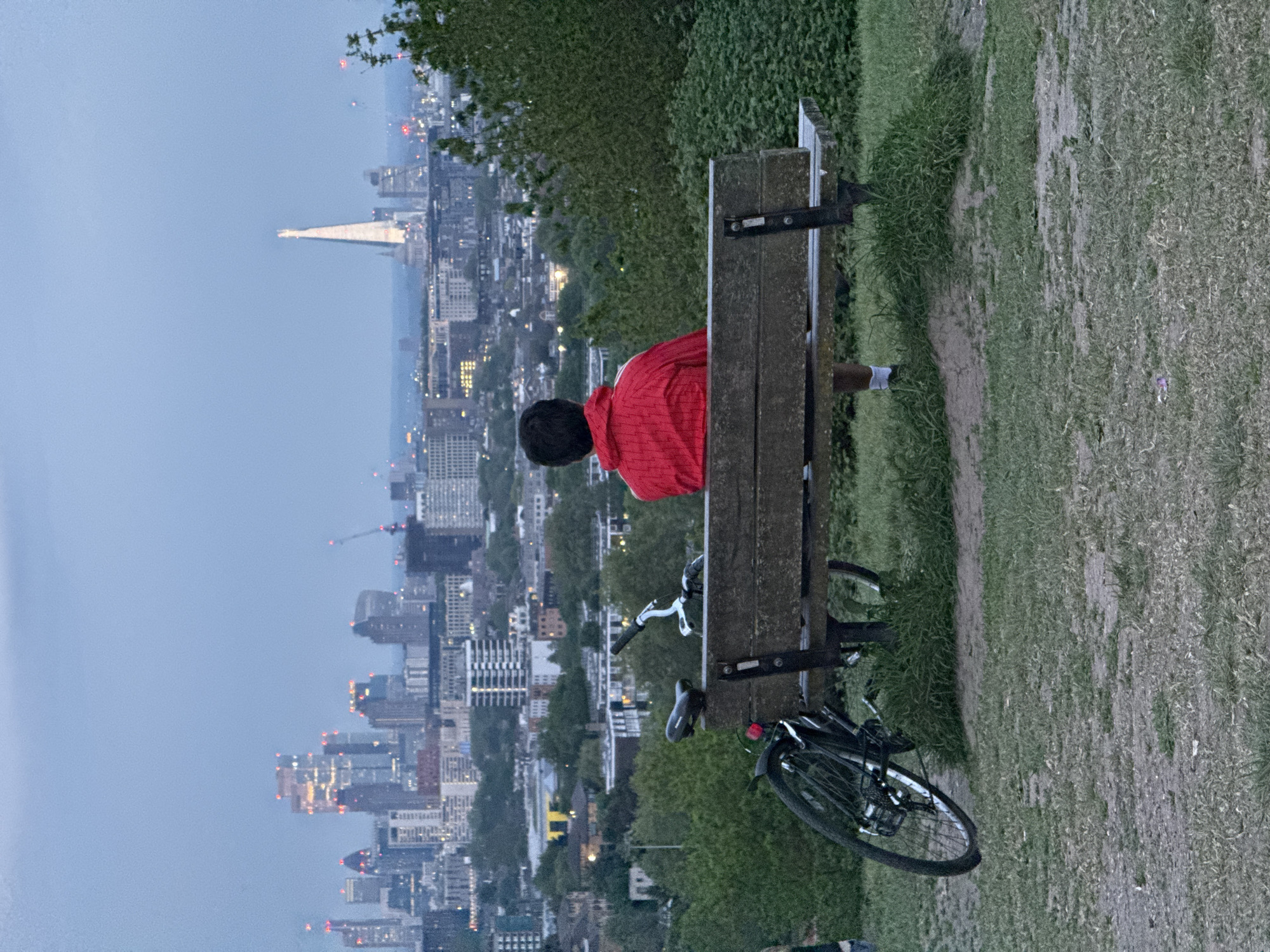 An individual wearing a red jacket is sitting on a park bench, with a bicycle leaning against the bench. The setting is a park overlooking a city, in London, with the Shard visible in the background. The time appears to be dusk, with city lights illuminated. The ground is covered in grass and a dirt path runs along the edge.