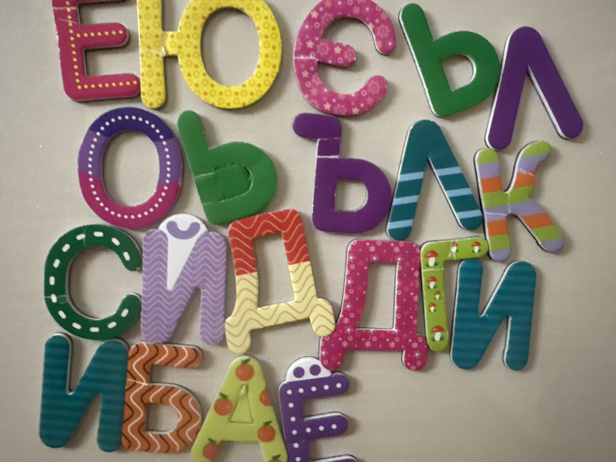 The image displays a collection of colorful, foam alphabet letters arranged on a neutral-colored, textured surface, likely a wall or a board. The letters are of various sizes and designs, some solid colored, others patterned with dots, flowers, stripes, or wavy lines. Some letters also have small illustrations, such as mushrooms or fruit. The letters appear to be magnetic or have a similar adhesive backing, suggesting they are intended for use on a magnetic surface. The arrangement of the letters is haphazard, with some overlapping. There are no people or animals visible in the image. The lighting is even, with no strong shadows, indicating an indoor setting. The image is a close-up, focusing on the letters themselves, with the background largely out of focus.
The provided location, Georgiyevsk, Russia, is not discernible from the visual information in the image. There are no landmarks, street signs, or other contextual clues that would allow for the identification of a specific geographic location. The scene depicted is generic and could be found in many indoor environments.