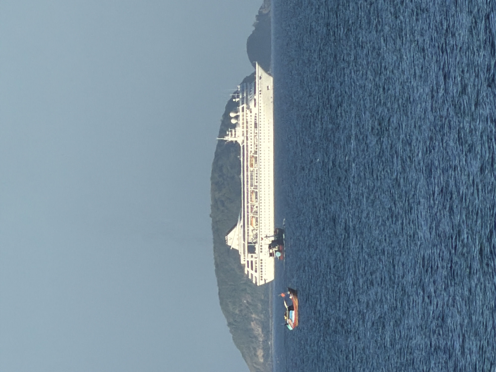 A wide shot captures a cruise ship and two smaller boats sailing on a calm blue sea. In the background, a lush green island rises from the water. The sky above is a clear, pale blue. This scene is characteristic of the waters off the coast of Nha Trang, Vietnam, a popular tourist destination known for its beautiful beaches and islands. The cruise ship, a large white vessel with multiple decks and windows, appears to be the main subject, while the two smaller boats, one a traditional wooden fishing boat and the other a more modern vessel, add a sense of scale and local context to the image.