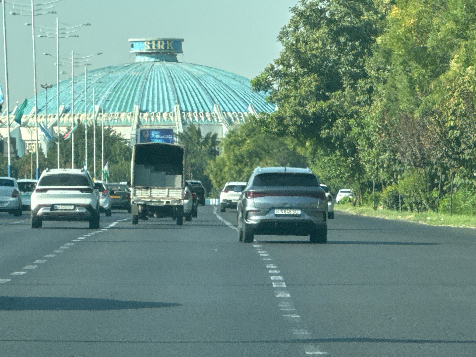 The image depicts a street scene in Tashkent, Uzbekistan, featuring a procession of vehicles moving along a multi-lane asphalt road. Dominating the background is the distinctive turquoise dome of the "Sirk" circus building, adorned with banners and flags. The road itself is marked with dashed white lines, indicating traffic lanes.
Several cars are visible in the foreground and middle ground, including a white Kia SUV on the left, a gray sedan on the right, and a white compact car further ahead. A truck with a flatbed trailer is positioned between the white Kia and the gray sedan. Trees and greenery line the right side of the road, suggesting an urban park or roadside vegetation. The sky is overcast, casting a diffused light on the scene. The overall impression is one of routine city traffic under a temperate sky, with the prominent circus building serving as a landmark.