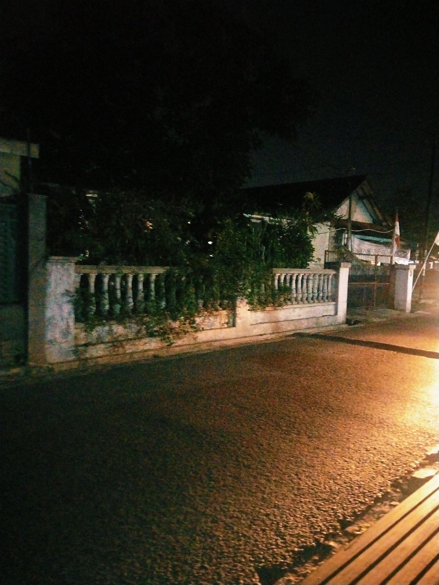 The image depicts a street scene at night, likely in Tasikmalaya, Indonesia, given the visible flag. A paved road occupies the foreground, illuminated by a bright light source from the right. To the left, a stone fence with ornate balusters runs along the property line of a house partially visible in the background. Overgrown vegetation climbs over the fence, partially obscuring the building. The house appears to be a modest structure with a sloped roof. A gate is visible in the background, and a flag, possibly the Indonesian flag, is planted on a pole to the right of the gate. The overall atmosphere is dim, with shadows and limited visibility due to the nighttime setting.