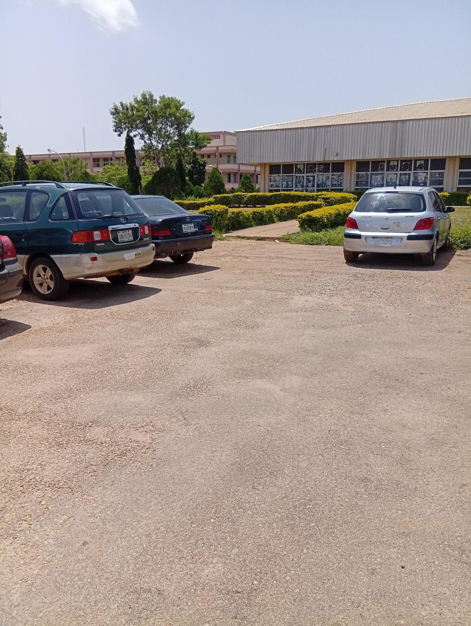 The image depicts a parking lot in front of a building, likely a university or administrative complex, in Gwong, Nigeria. The building has a long, horizontal design with a light-colored, corrugated roof and numerous windows on the facade. Yellow flowering bushes line the front of the building, creating a hedgerow effect. Several cars are parked in the lot, which is covered in gravel.
In the foreground, to the left, is the rear of a dark-colored vehicle, partially visible. Next to it is a dark green SUV, identified by the "TOYOTA" and "Pacific" badges on its rear. This SUV has a pale beige lower body section and its rear bumper is visible. To the right of the SUV is a dark-colored sedan, possibly a Mercedes-Benz based on the emblem visible on its rear. Further to the right and slightly more distant is a silver hatchback, identified as a Peugeot 307 by the badge on its trunk. All vehicles are parked and appear stationary.
The environment suggests a dry, sunny day with a clear blue sky and a few wispy clouds. Trees, including a tall, slender one and broader, leafy ones, are scattered around the scene, adding to the sense of a campus or institutional setting. The overall impression is one of an orderly, daytime scene with no immediate signs of activity or disturbance.