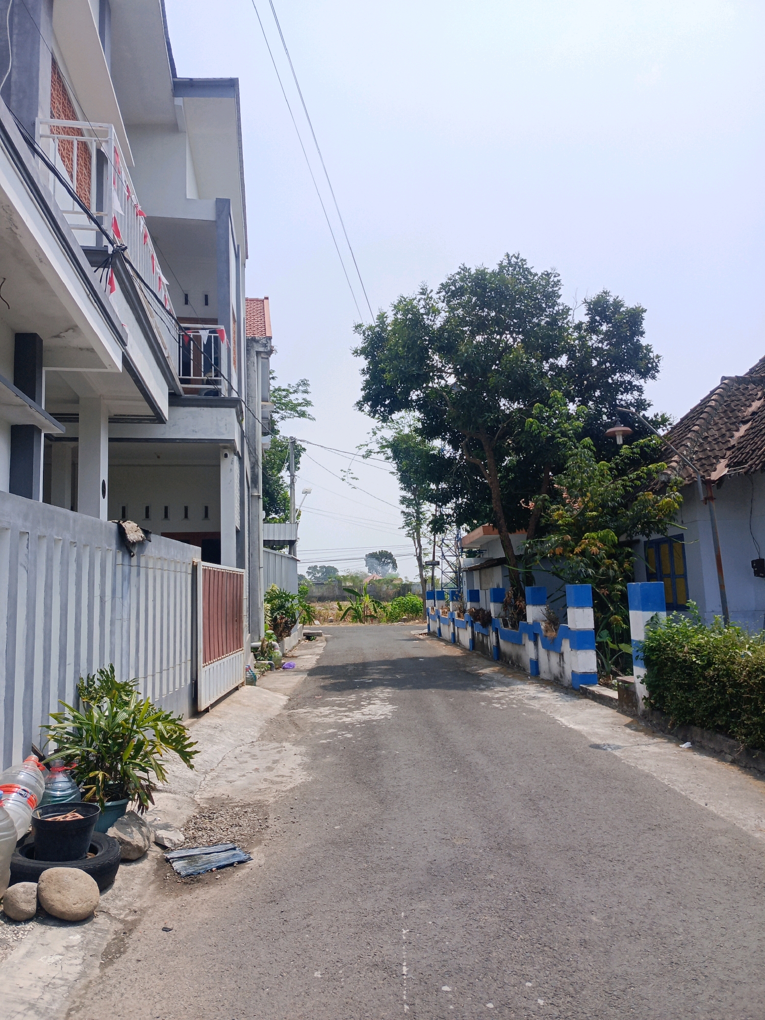The image shows a narrow asphalt road in the Mangkujayan area of Indonesia, bordered by residential buildings and vegetation. On the left, a modern multi-story building with a grey and white facade is visible, featuring balconies decorated with small Indonesian flags. A white fence with a brown gate separates the property from the street. Potted plants, discarded tires, and rocks are present on the sidewalk in front of the fence.
On the right side of the road, a traditional-looking house with a tiled roof and blue and white decorative pillars along its perimeter is seen. A large tree with lush green foliage stands between the two properties, partially obscuring a street lamp. Beyond the immediate residential area, the road continues into the distance, with more houses and vegetation visible under a clear sky. Electrical wires are strung across the scene.