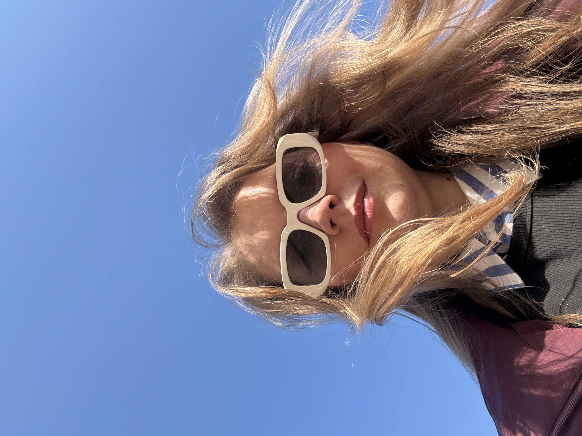 A woman with long blond hair wearing sunglasses stands outdoors under a clear blue sky. She is wearing a striped shirt under a black sweater and a burgundy jacket. The photograph is taken from a low angle, looking up at the woman. The location is not discernible from the image.
