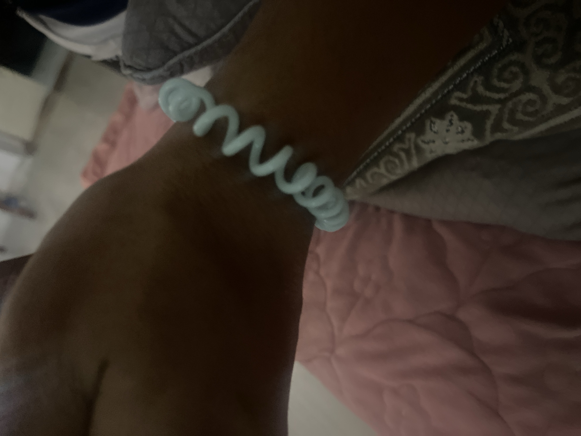A close-up shot focuses on a person's wrist wearing a light blue, spiral-shaped bracelet. The bracelet is coiled around the wrist, with its translucent plastic material clearly visible. The skin tone of the wearer appears to be medium brown.
In the background, a textured pink surface, possibly a blanket or comforter, is visible. Above that, a patterned dark gray fabric with swirling designs can be seen, suggesting clothing or upholstery. The lighting appears to be indoor and somewhat dim, creating soft shadows.
The image is likely taken indoors, with no external environmental cues like time of day or weather being discernible. The focus is entirely on the bracelet and the immediate surrounding area of the arm. No text is visible in the image.
