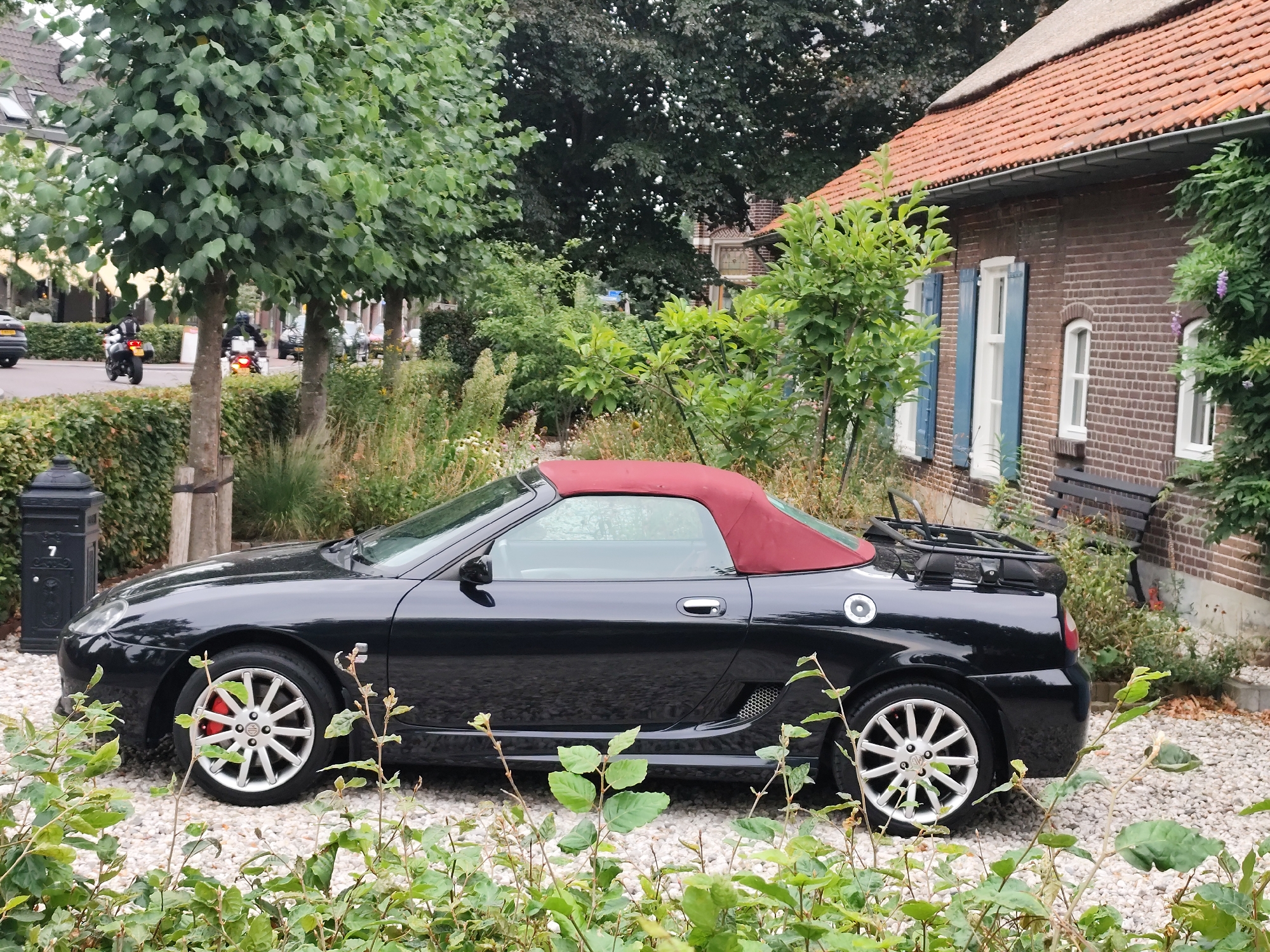 A black MG TF convertible with a red soft top is parked on a gravel driveway in front of a brick house with an orange tile roof. The car is angled towards the left side of the frame, with its front wheel visible. The house has blue shutters on its windows. In the background, there are trees and other vegetation. To the left of the car, a black lamppost with a number "7" is visible. Further in the background, on a paved road, two motorcyclists are riding. The environment suggests a suburban or rural setting. The image appears to have been taken in Schaijk, Netherlands.