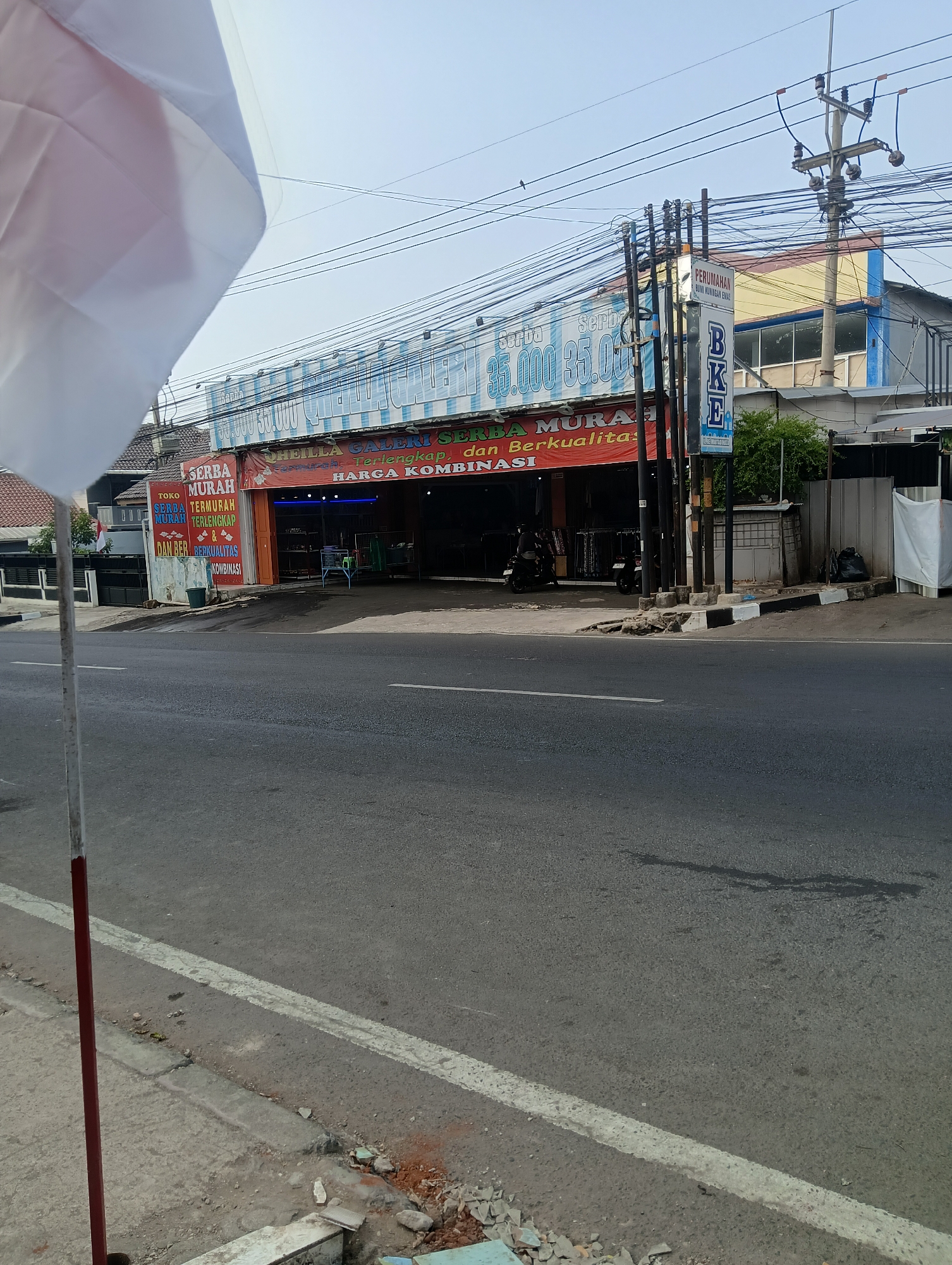 The image shows a street scene in Ciporang, Indonesia. In the foreground, a portion of a white flag on a red and white pole is visible on the left side. The street itself is asphalt with a white line marking the lane. On the right side of the street, a building with several signs is prominent. One sign displays "SERBA MURAH" (Very Cheap) and "SERBA TERMUHUR MURAH & DAN BERKUALITAS" (Very Cheap, Cheapest, Cheap & Quality). Another large banner above the entrance of the building advertises "SHEILLA GALERI SERBA LENGKAP" (Sheilla Gallery Complete) and "HARGA KOMBINASI" (Combination Price). The building appears to be a shop or store, with the entrance open, revealing shelves and items inside. A few motorcycles are parked near the entrance, and two individuals are visible in the background, one on a motorcycle. Further down the street, a utility pole with numerous wires is situated in front of another building. The sky is clear and bright.
