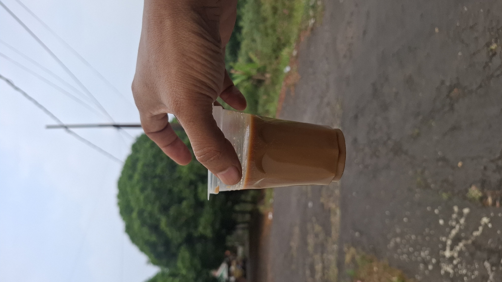 A hand is holding a clear plastic cup filled with a brown liquid, likely coffee, outdoors. The cup is held from the side, with the thumb on top and fingers wrapped around the bottom. The background is slightly blurred, showing a paved road, some greenery, and a large, rounded tree. Overhead, power lines are visible against a cloudy sky. The scene appears to be in Cilaku, Indonesia, given the context.