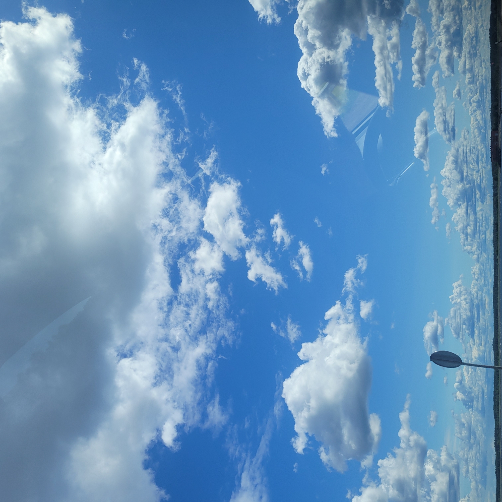 The image depicts a bright, clear blue sky filled with a variety of clouds, ranging from large, puffy cumulus clouds to wispy cirrus formations. The scene is viewed from what appears to be ground level, looking upwards and towards the horizon. In the foreground, a solitary, grey traffic sign with an oval-shaped head stands on a thin pole, angled slightly to the right. The horizon line is visible at the bottom of the frame, suggesting a body of water such as a lake or river, bordered by a distant, dark treeline. The reflection of the sky and clouds is subtly visible on a surface in the upper part of the image, possibly a window. The overall impression is one of a calm, expansive day. The location is Odukalns, Latvia.