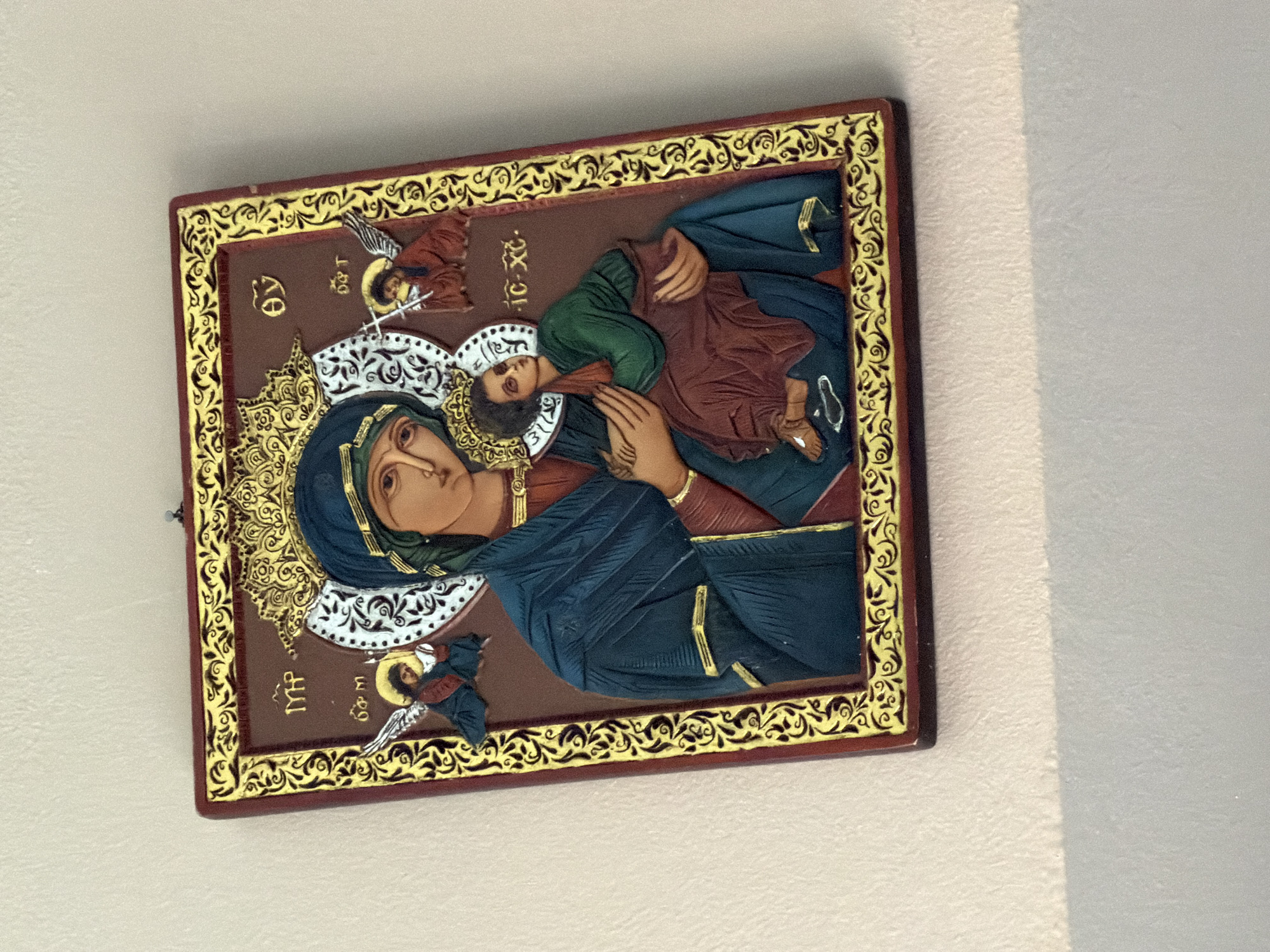 This is a religious icon depicting the Virgin Mary holding the infant Jesus. The icon is framed with a decorative border. The Virgin Mary wears a blue robe and a veil, with gold ornamentation around her head. The infant Jesus is held in her arms, dressed in a green tunic. Two small angels are depicted flanking the central figures. The background of the icon is a deep brown, with decorative elements in gold. There are also some Cyrillic inscriptions on the icon, though they are difficult to read clearly. The icon is mounted on a wall, suggesting it is a religious artifact displayed in a home or place of worship. The lighting suggests it is indoors, possibly during the daytime, as there is natural light casting shadows.