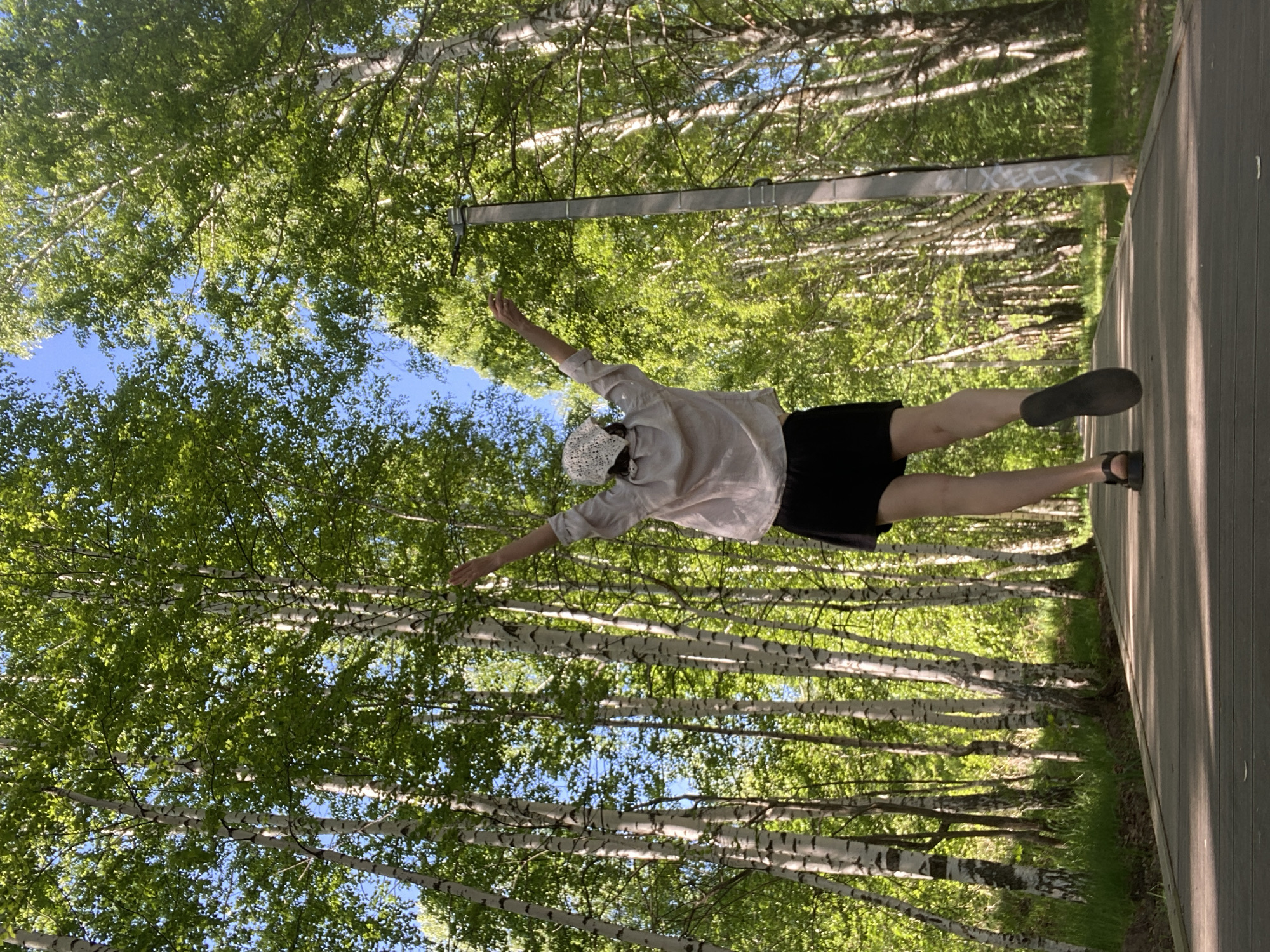 A person lies on a wooden deck amid a birch forest. The scene is oriented at an angle. The person is wearing a light-colored shirt, a black skirt, and a white head covering. Their arms are outstretched, appearing to touch the surrounding trees. The forest consists primarily of birch trees with green leaves, visible against a backdrop of blue sky. The setting is located in the city of Chaikovsky, Russia. A grey pole and graffiti are visible behind the person.